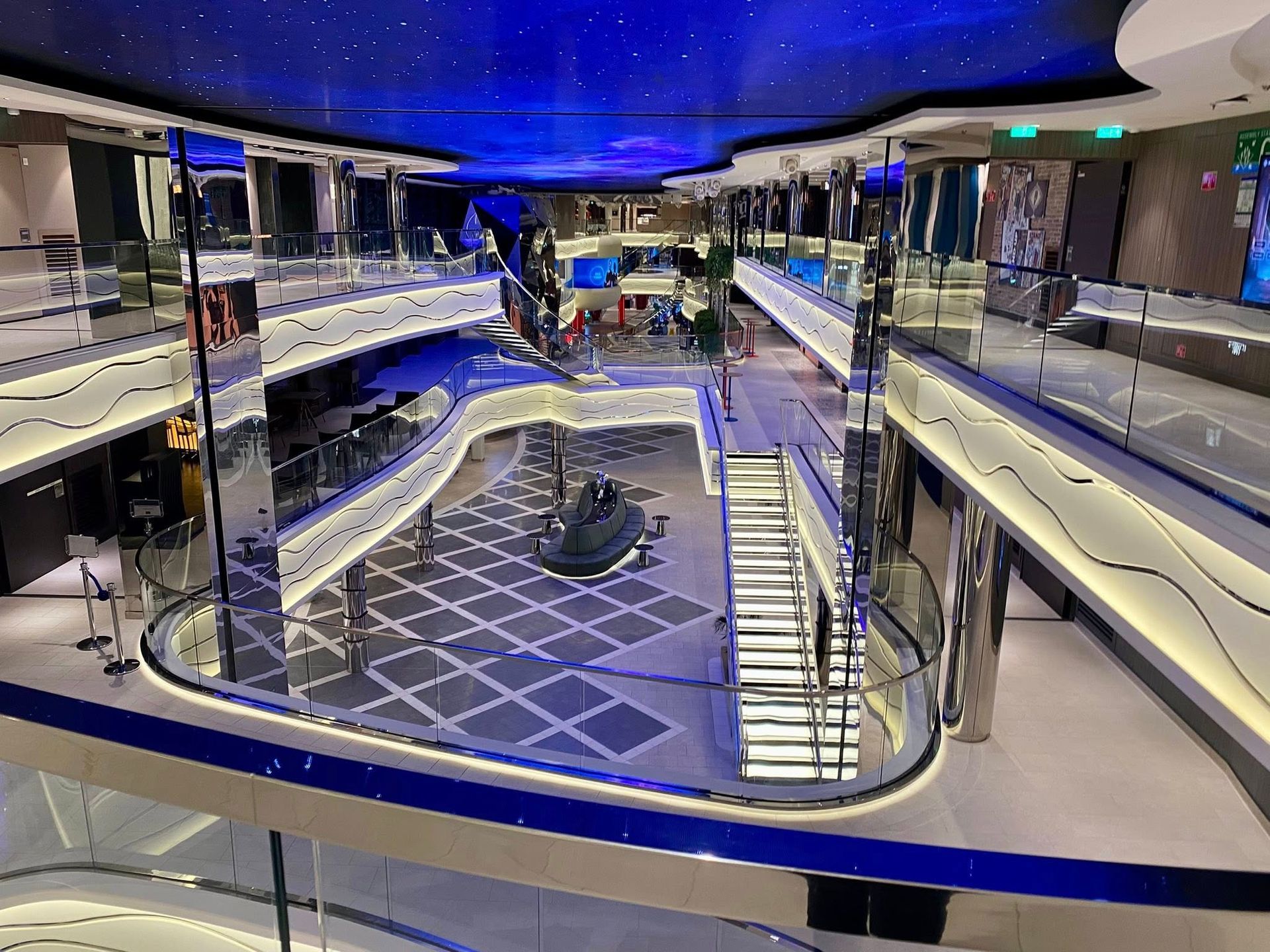 Multi-level cruise ship atrium with white railings, a tiled floor, and a blue, star-patterned ceiling with ambient lights.