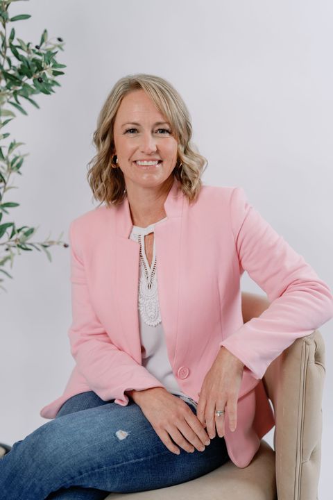 A smiling person sits on a chair wearing a light pink blazer and blue jeans, with a small potted tree in the background.