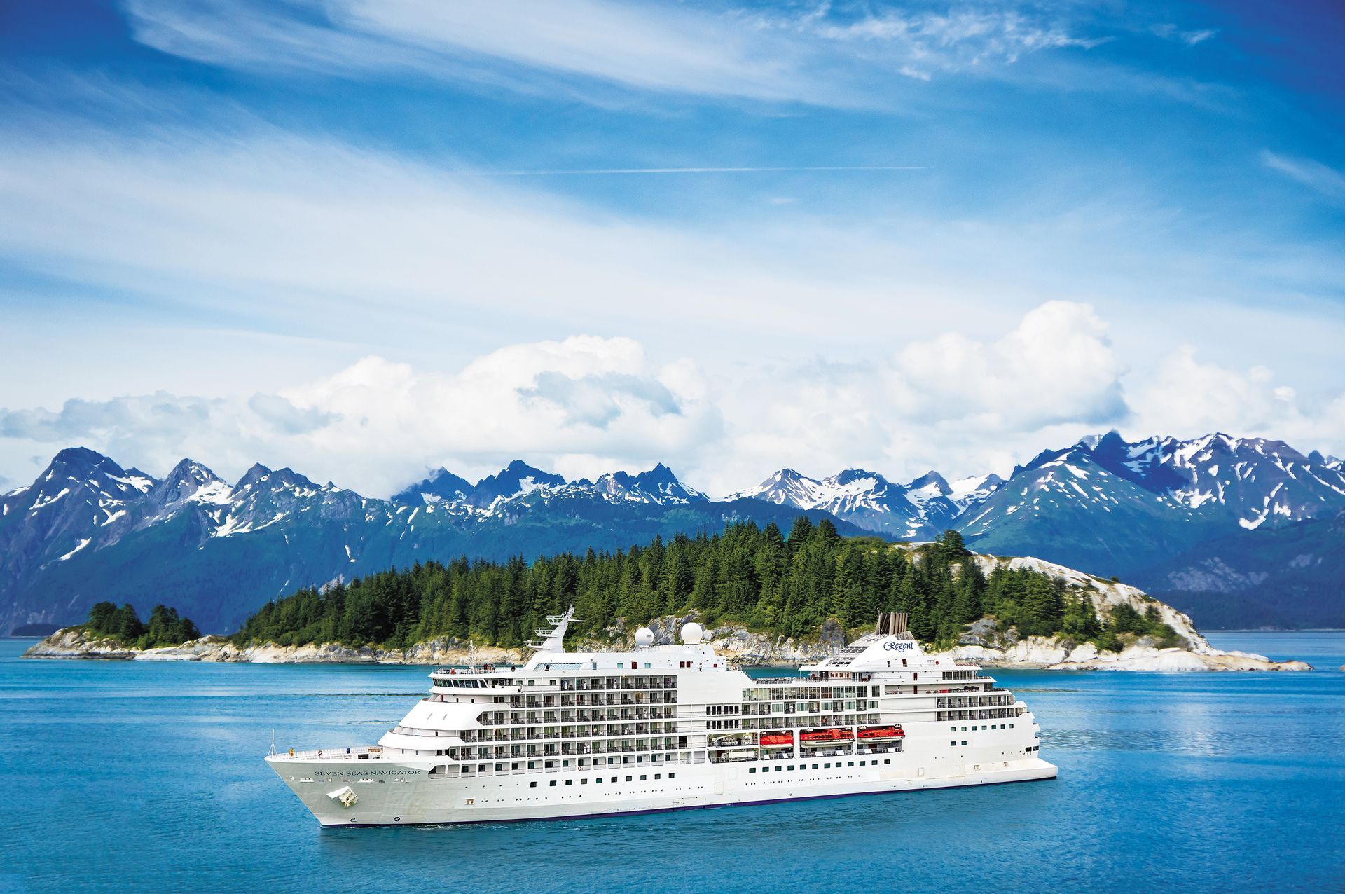 A white cruise ship sails across blue water past a forested island with snow-capped mountains in the background.