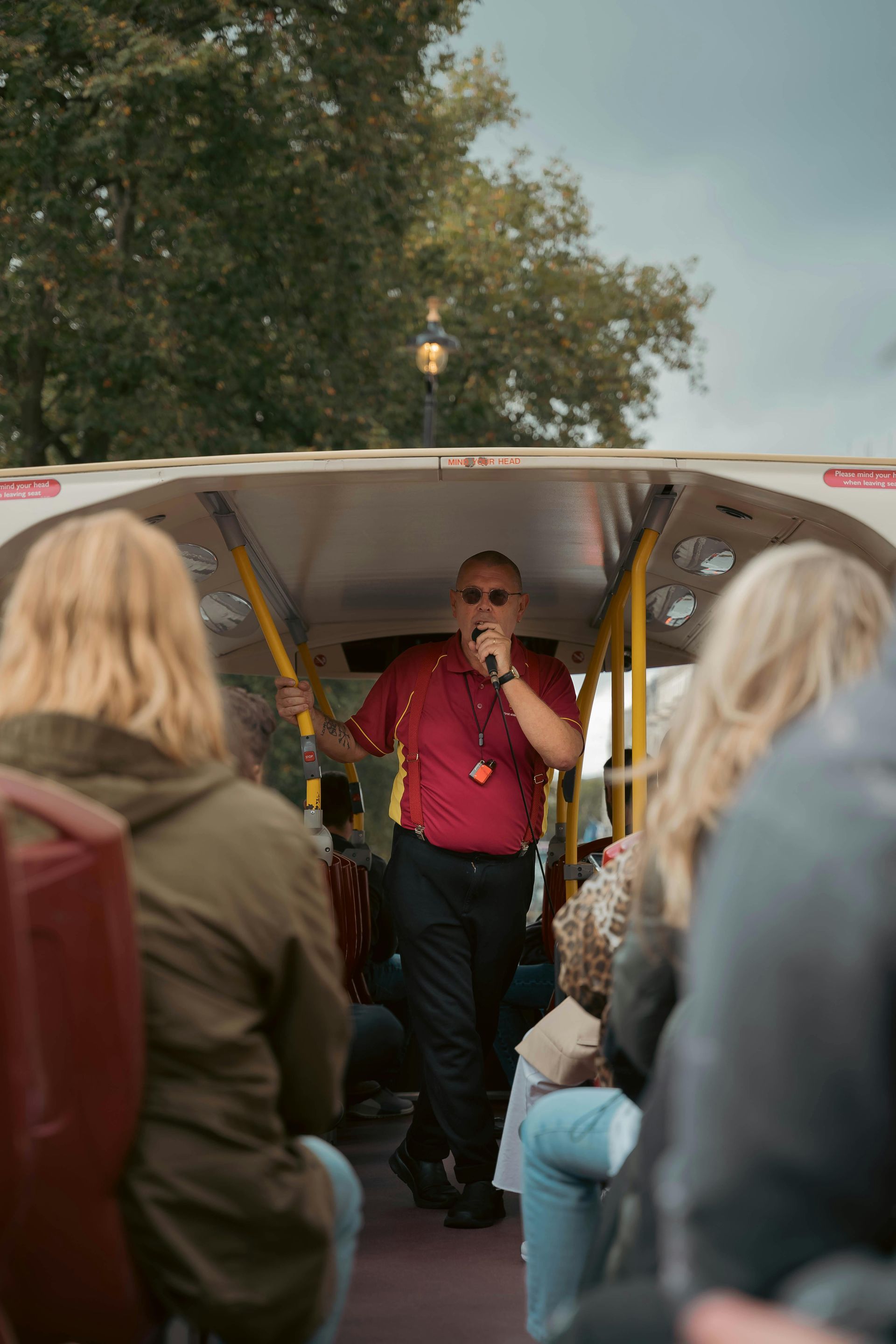 A tour guide in a red shirt speaks into a microphone while standing on the upper deck of an open-top bus.
