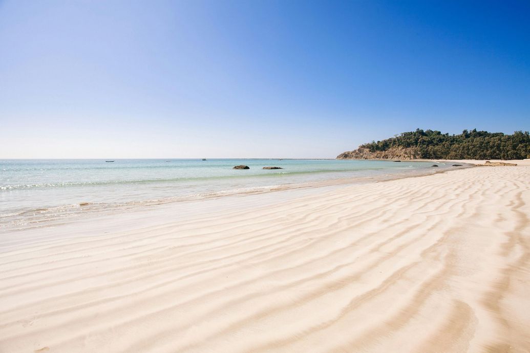 A bright, sunny beach with gentle wave patterns in the sand leading to calm, light blue water and a distant, tree-lined hill.