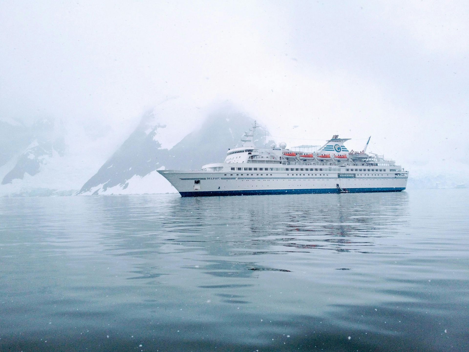 A white cruise ship floats in calm, gray water in front of a misty, snow-covered mountain range.