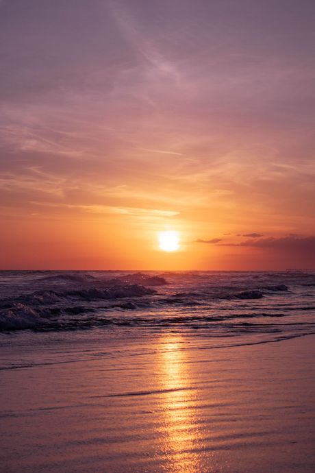 A vibrant sunset over a calm ocean, casting a golden reflection on the wet sand at the shoreline.