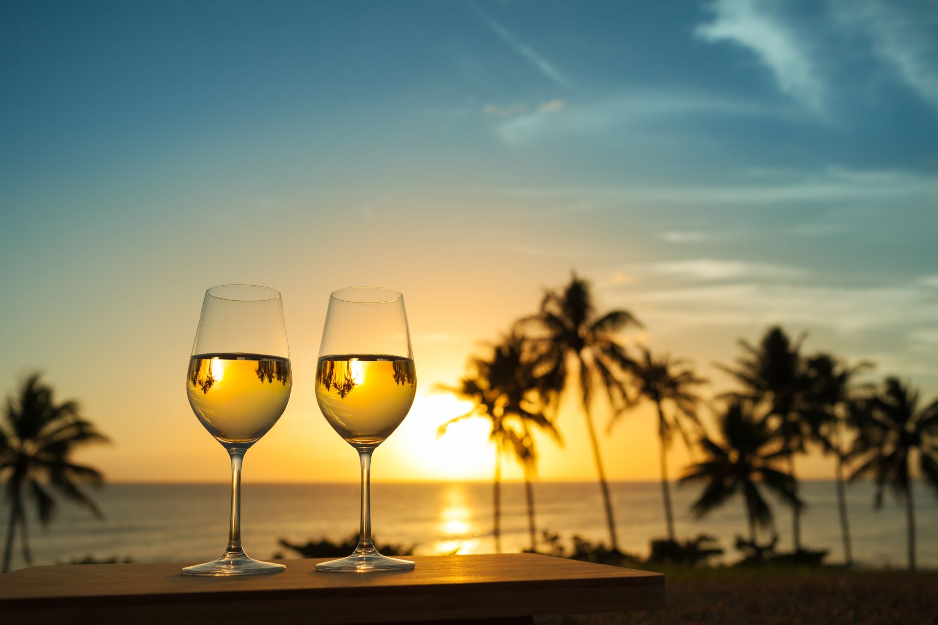 Two wine glasses filled with white wine stand on a ledge overlooking a tropical beach at sunset with palm trees.