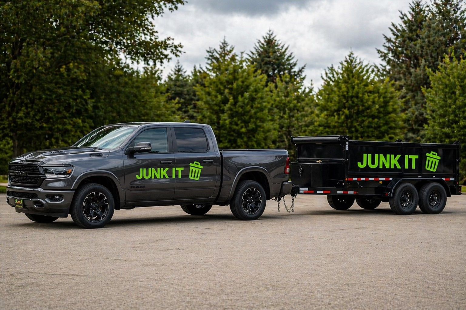 Black junk removal pickup truck towing a trailer with neon green “JUNK IT” lettering on a road, trees behind.