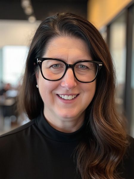 Headshot of a smiling person wearing black-rimmed glasses and a black mock-neck top in an office setting.