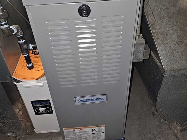 Silver furnace cabinet with vent slots and stickers in a utility closet, beside an orange pipe/valve.