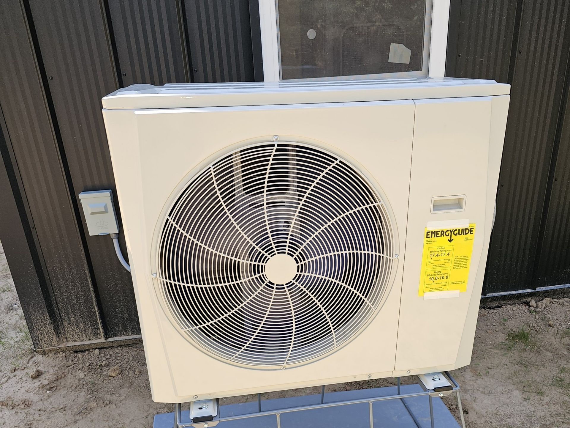 Outdoor beige air-conditioning unit with a large fan, mounted beside a wall with a yellow service tag.