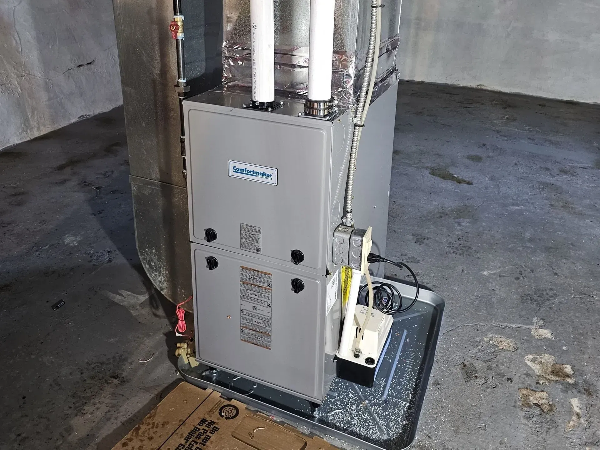 Gray furnace unit in a basement utility area beside exposed pipes and a dark floor.