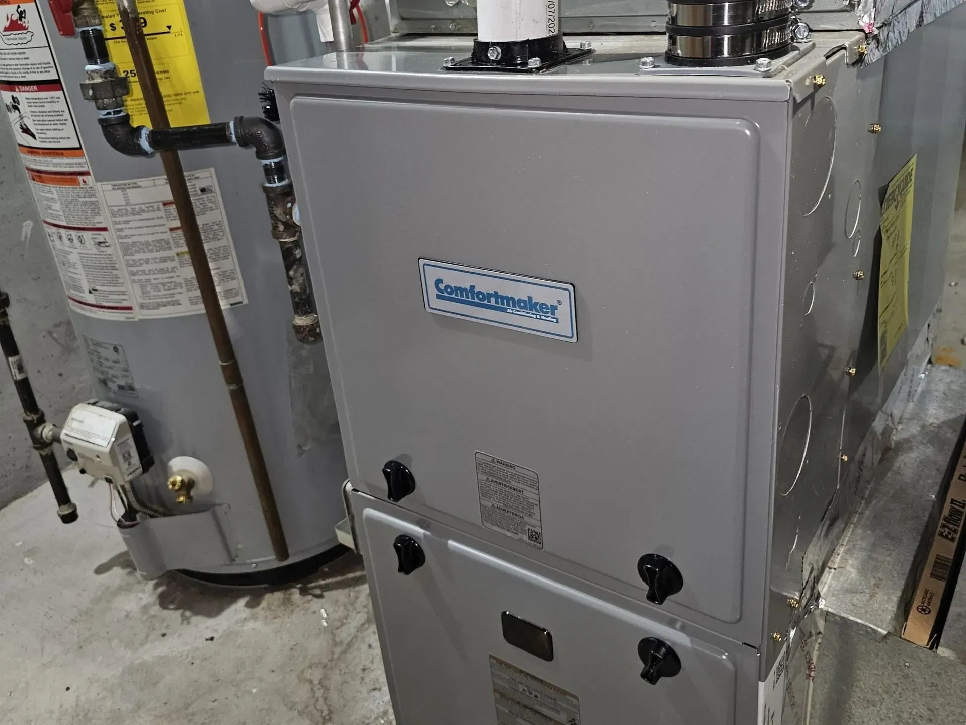 Gray furnace in a utility room with pipes and vents, viewed from the front.