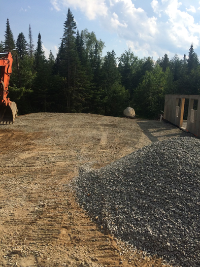 Construction site with excavator, gravel pile, and dirt road beside trees and a small building