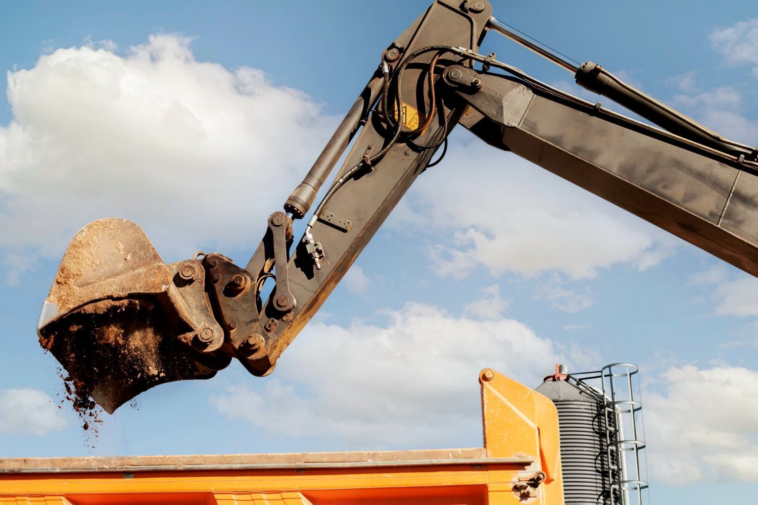 Excavator bucket dumping soil into an orange truck against a blue sky