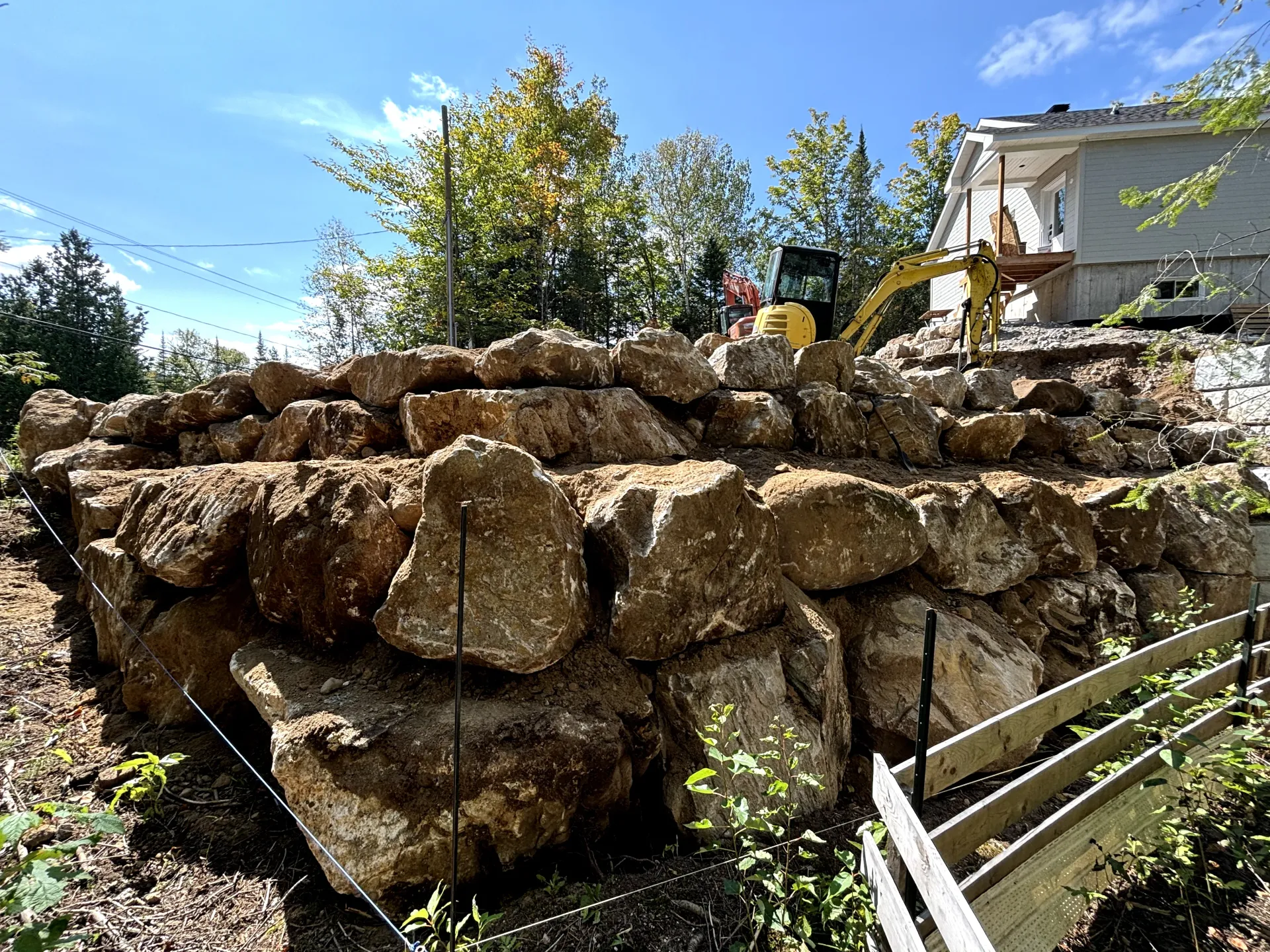 Construction site with gravel pad, white drainage pipes, and an excavator near a wooded area