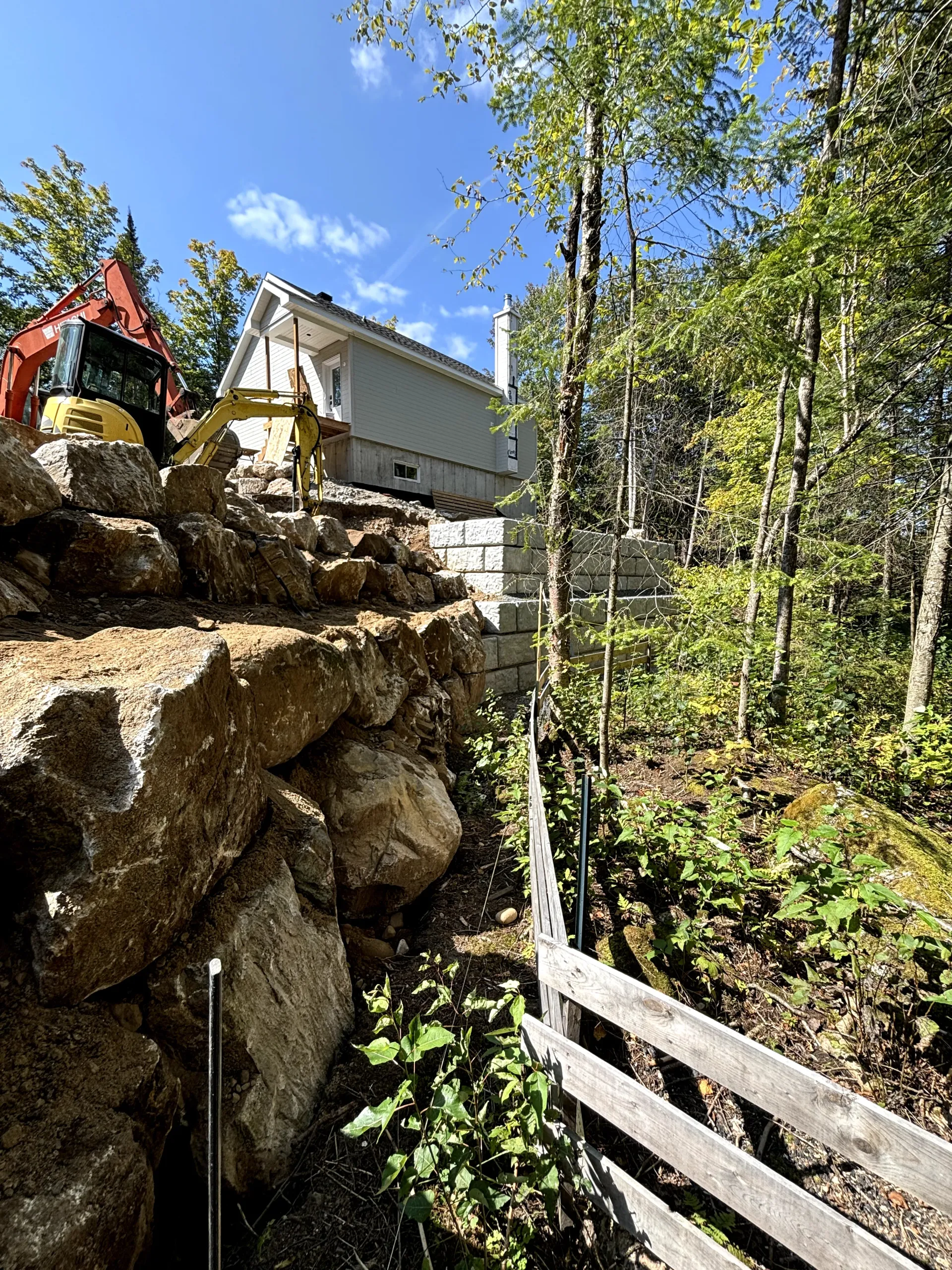 Rocky hillside with trees, a wooden fence, and a white house under a blue sky