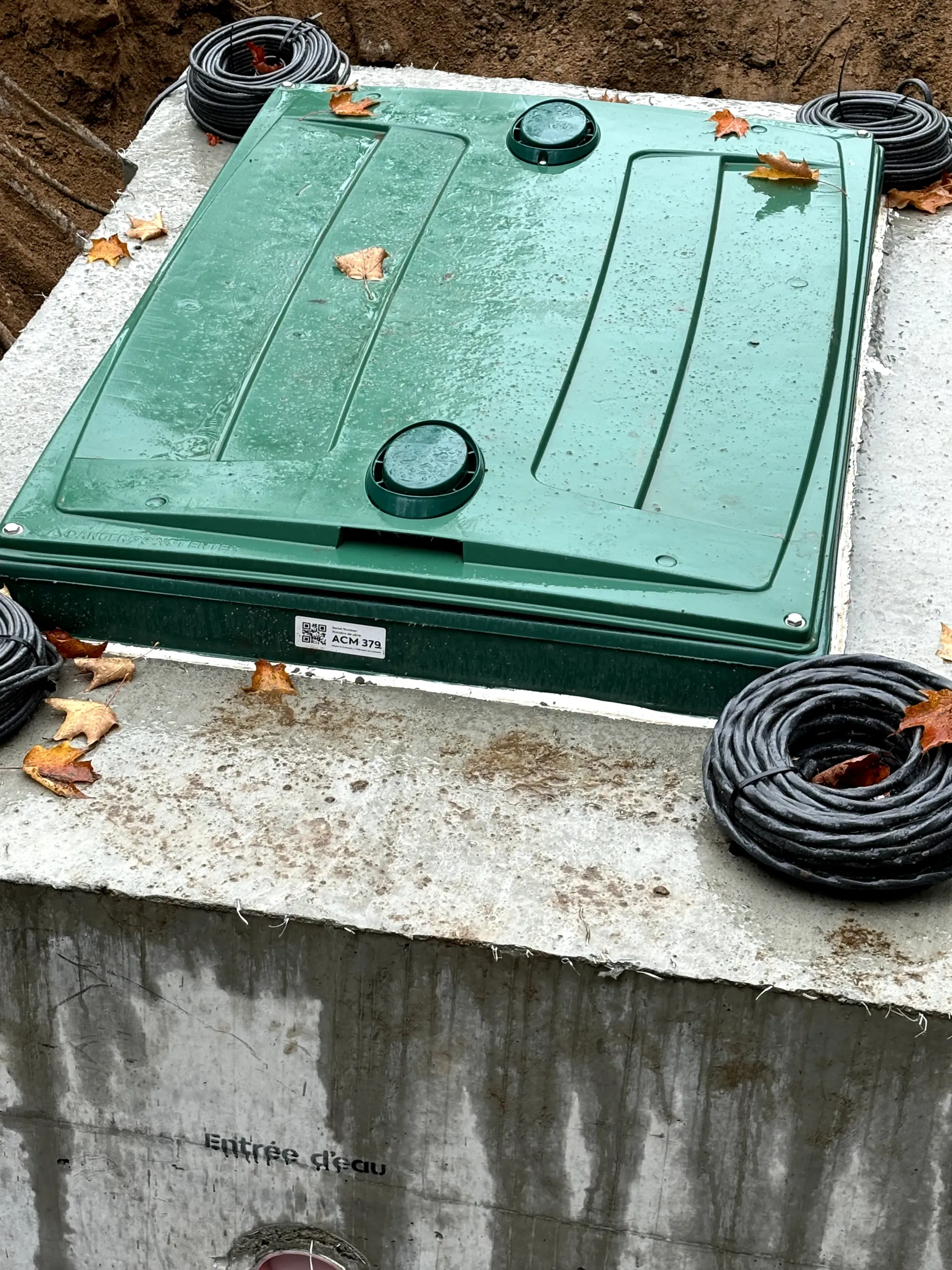 Green utility hatch on a concrete surface, surrounded by coiled black cables and fallen leaves.