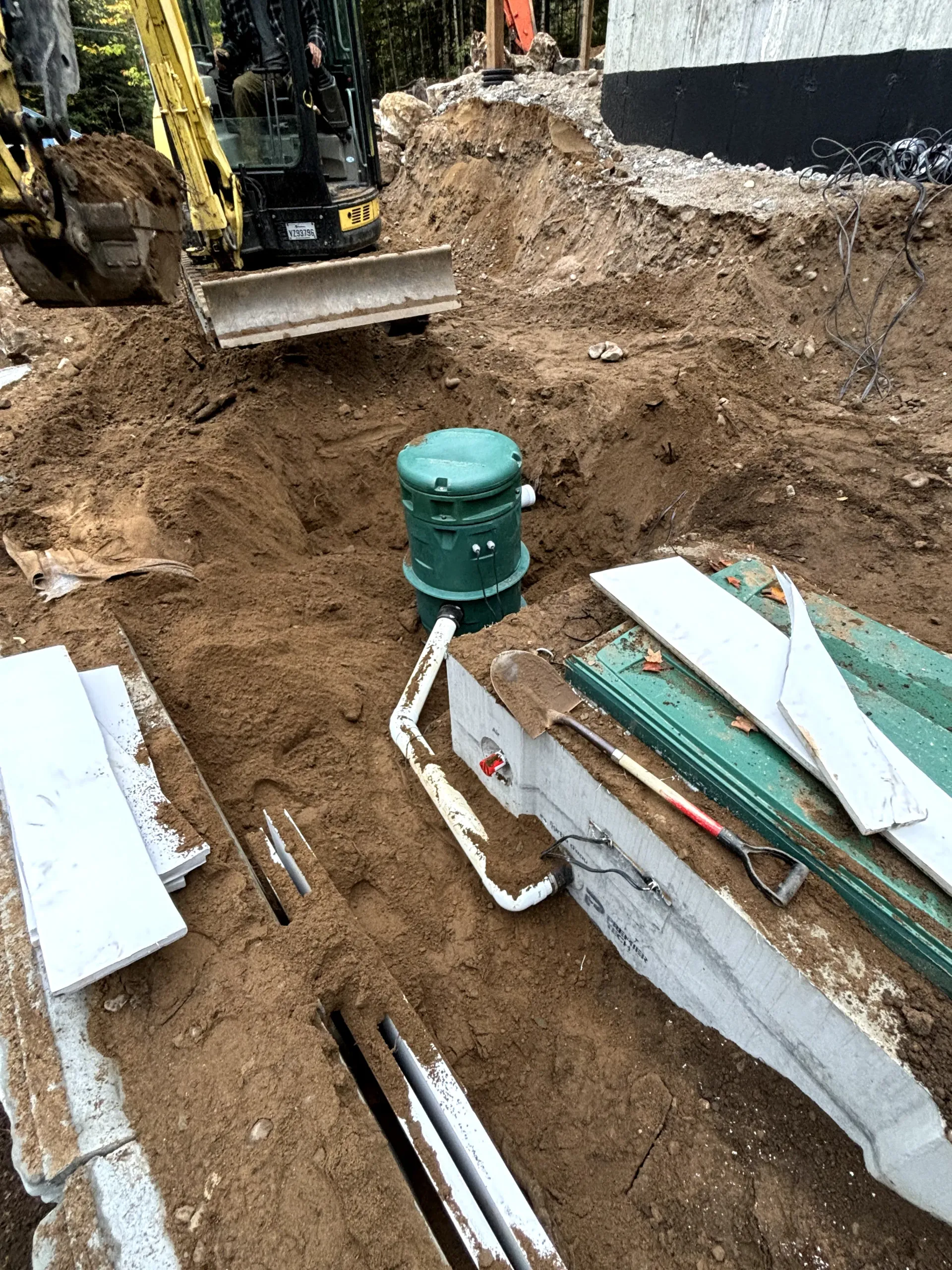 Construction trench with green utility cover, white pipes, and excavator near exposed dirt and concrete slabs