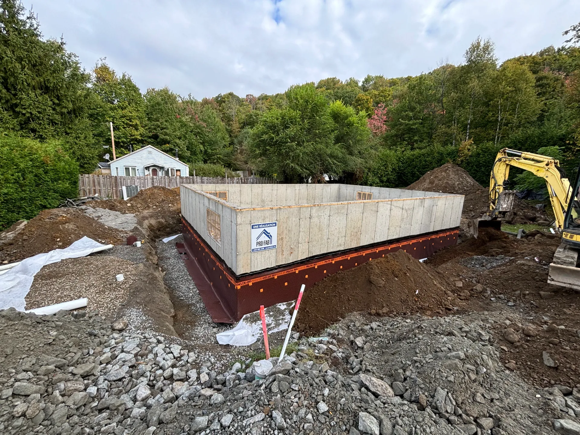 Construction site with concrete foundation walls, excavator, and gravel in a wooded area