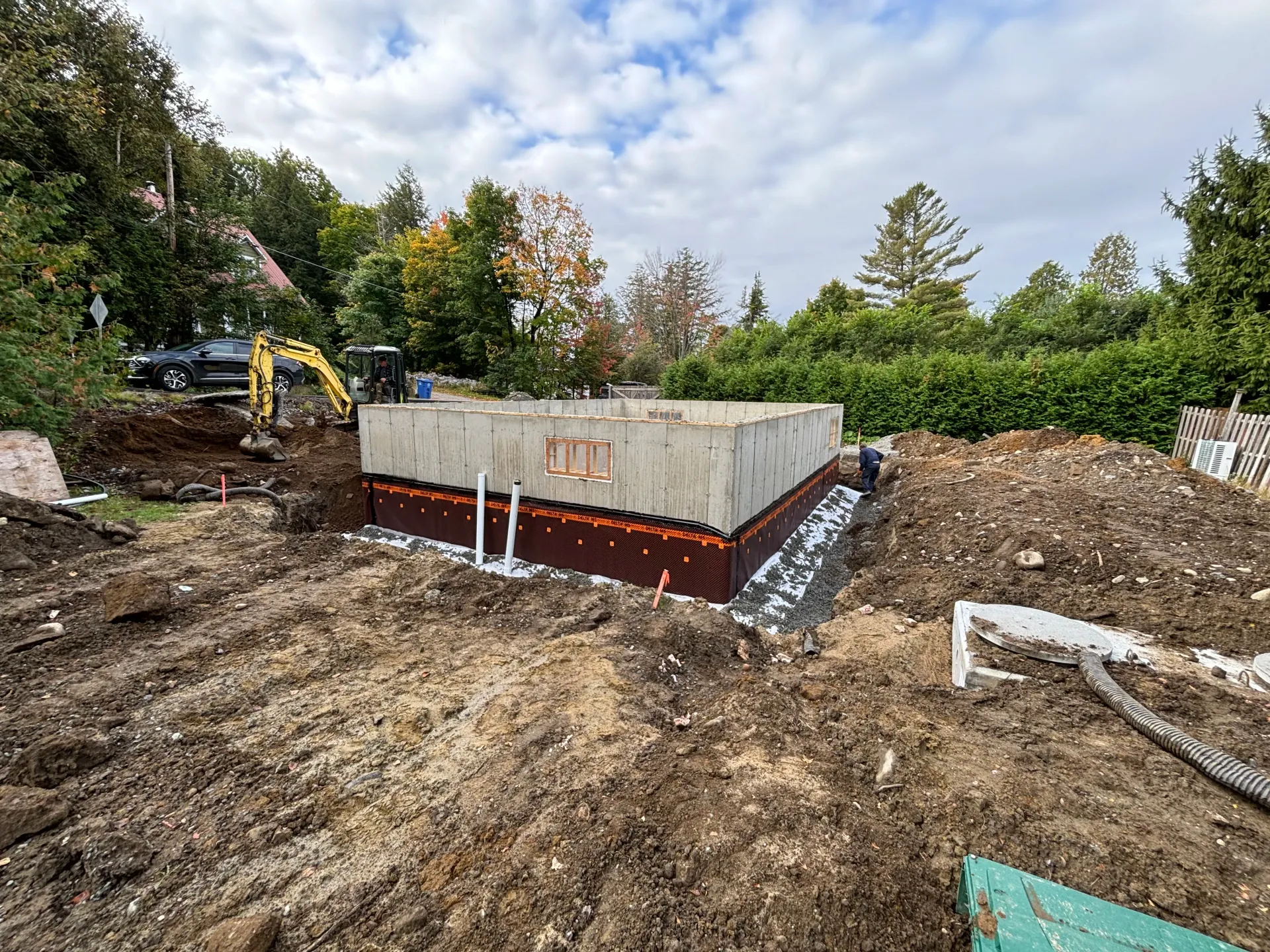 Construction site with concrete foundation walls, excavator, dirt piles, and trees under a cloudy sky