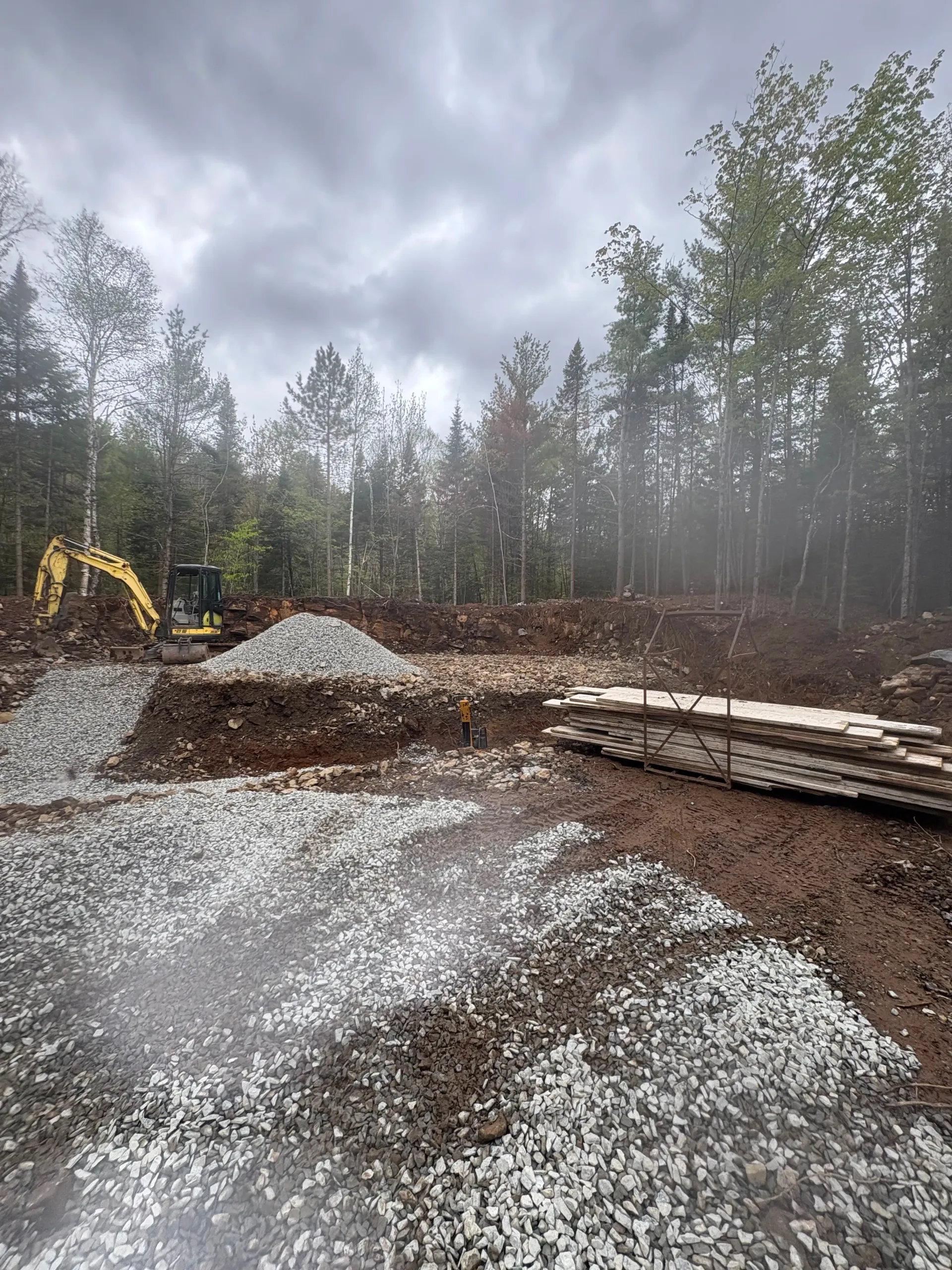 Gravel piles and a yellow excavator in a muddy wooded clearing under a cloudy sky
