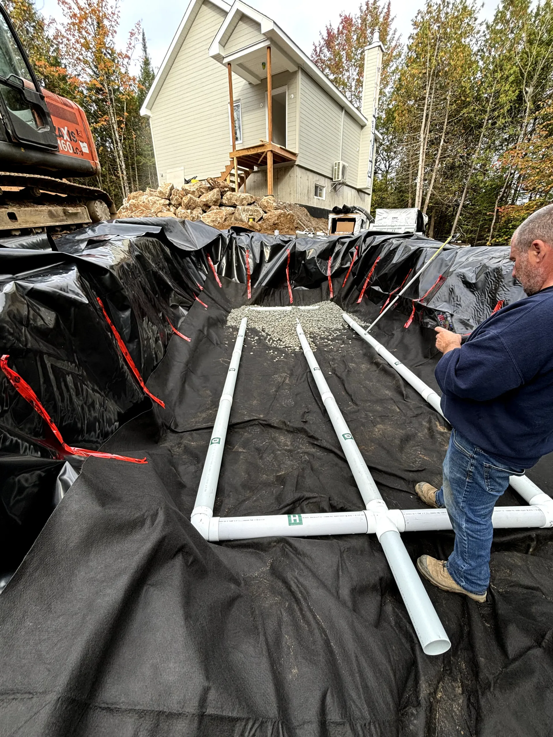 Person standing by a black-lined backyard excavation with white PVC pipes and a house in the background