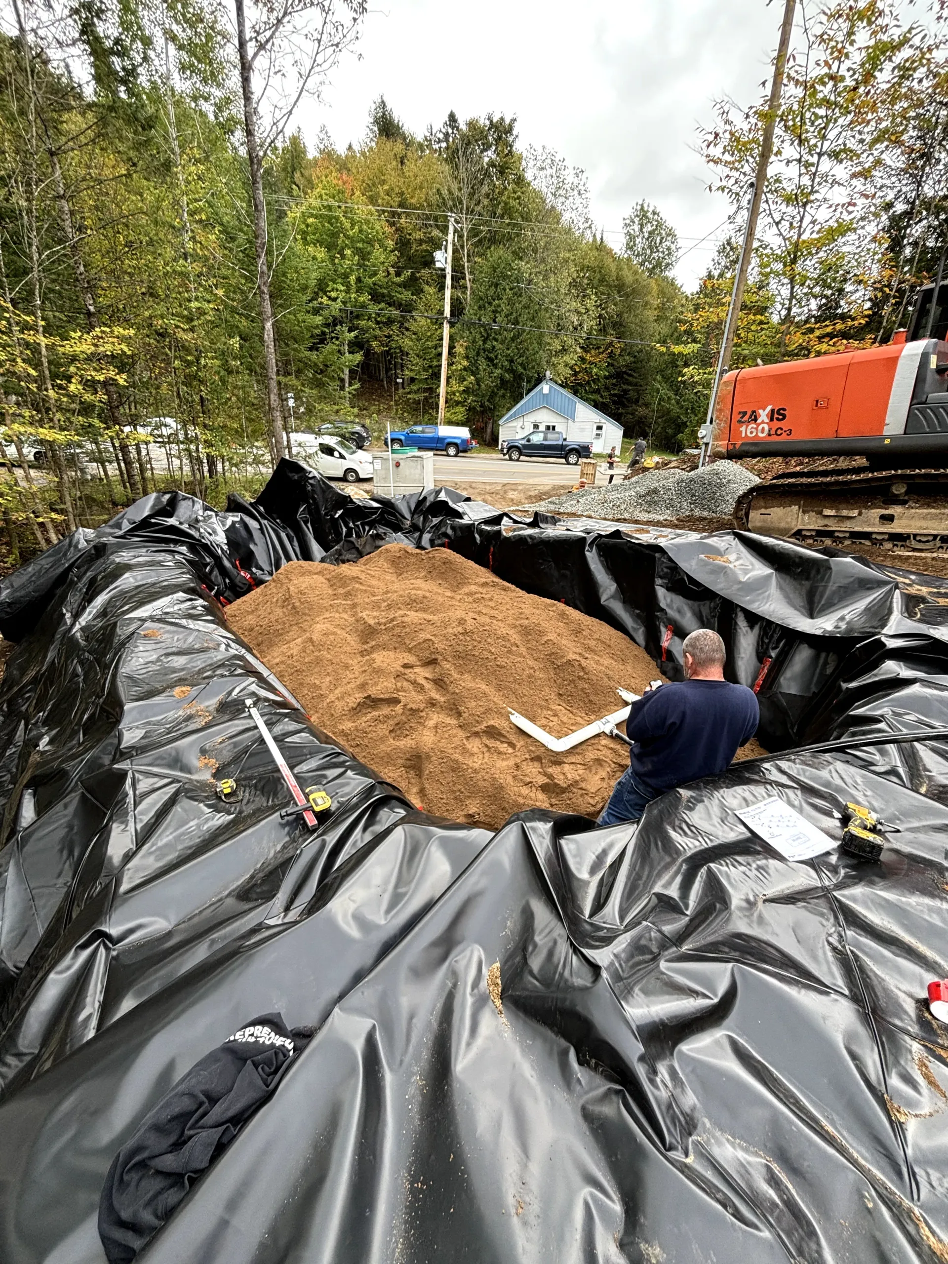 Worker spreading soil in a black-lined excavation beside trees and a red truck
