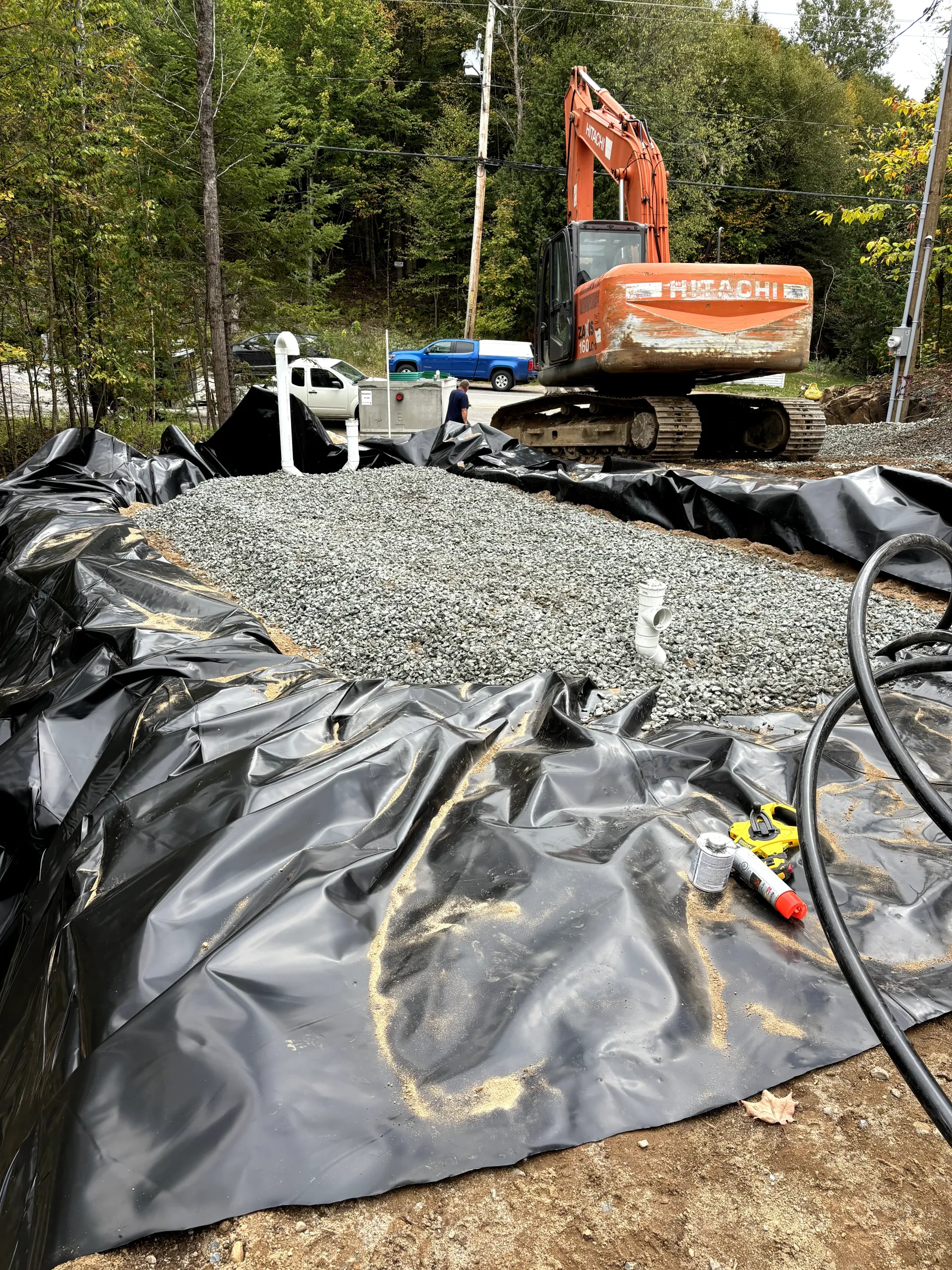 Excavator on black tarp spread over gravel at a wooded construction site.