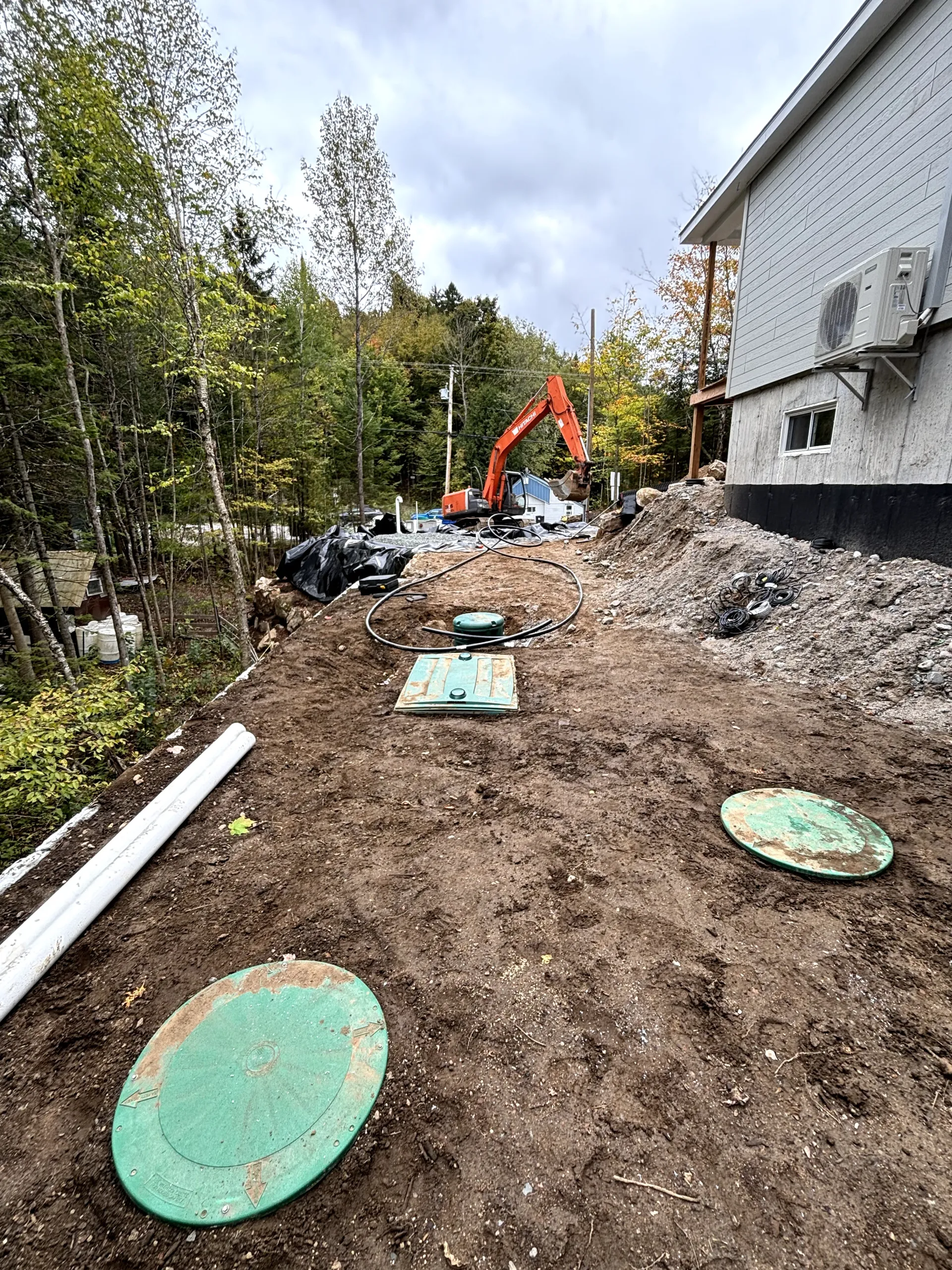 Construction site beside house with excavator, dirt path, and green utility covers
