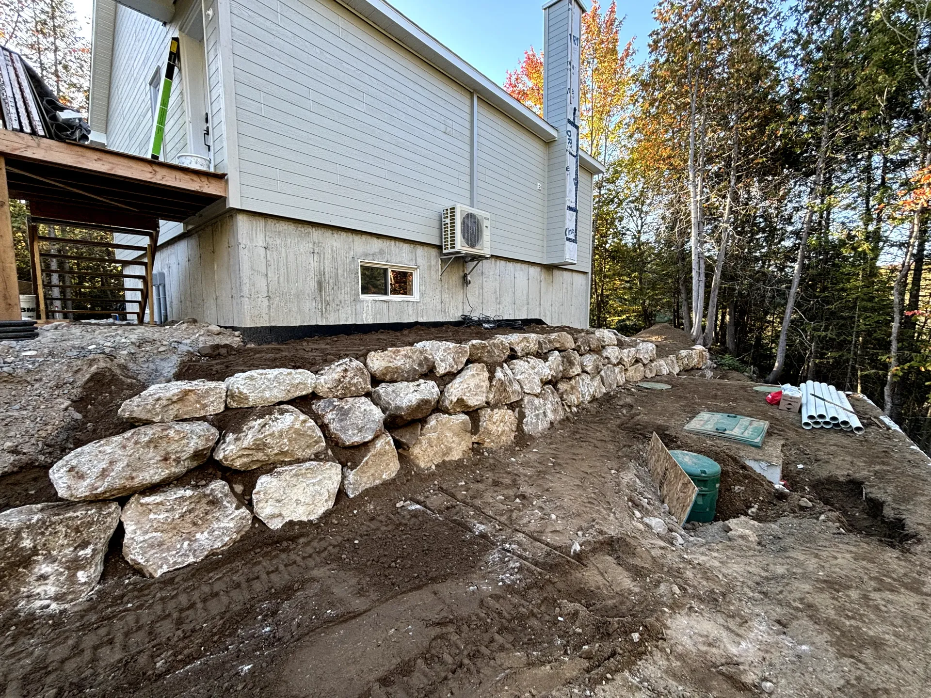 Stone retaining wall beneath a white house on a dirt slope, with trees and construction materials nearby