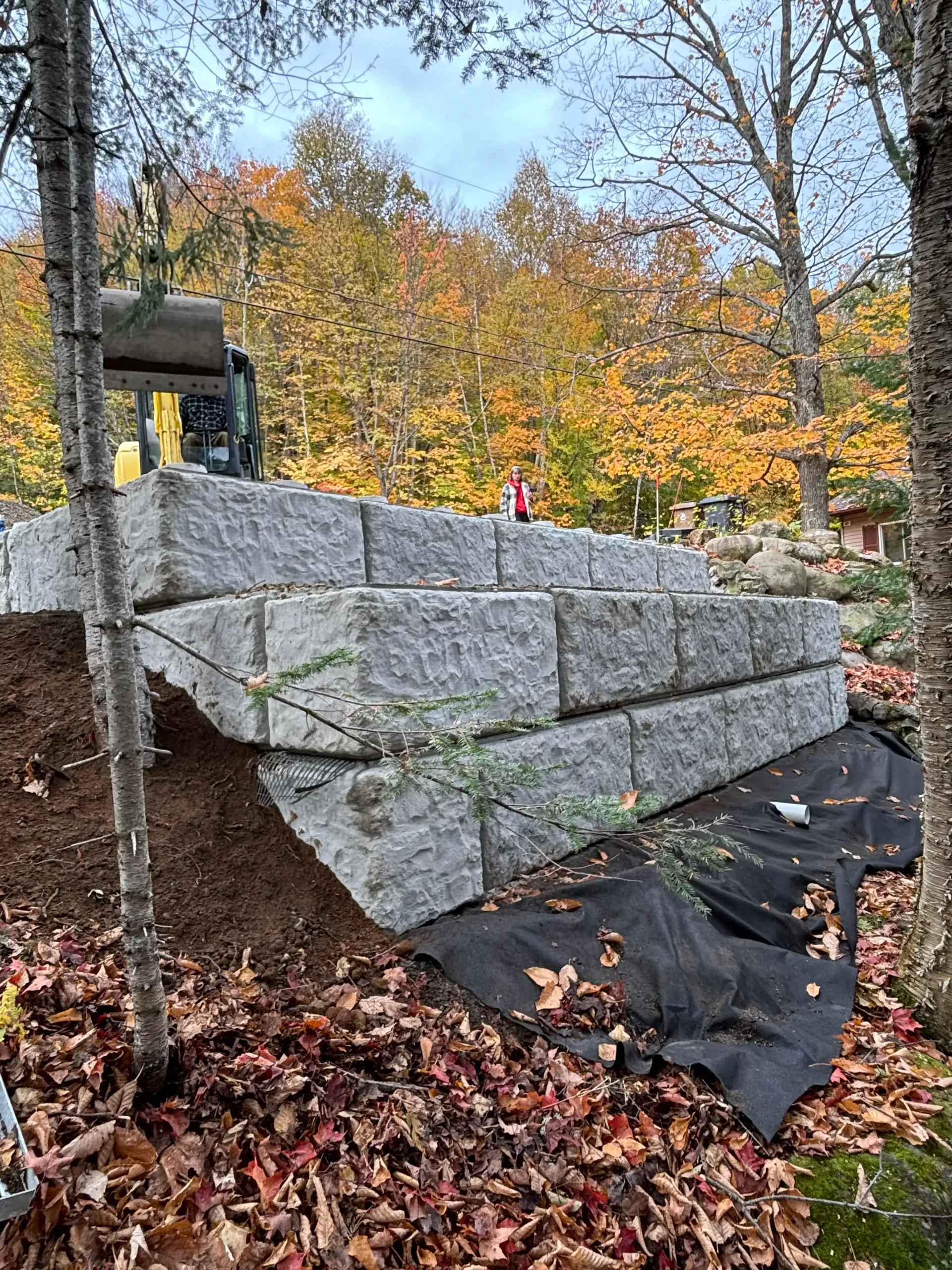 Stacked gray concrete blocks form a retaining wall at a wooded construction site in autumn.