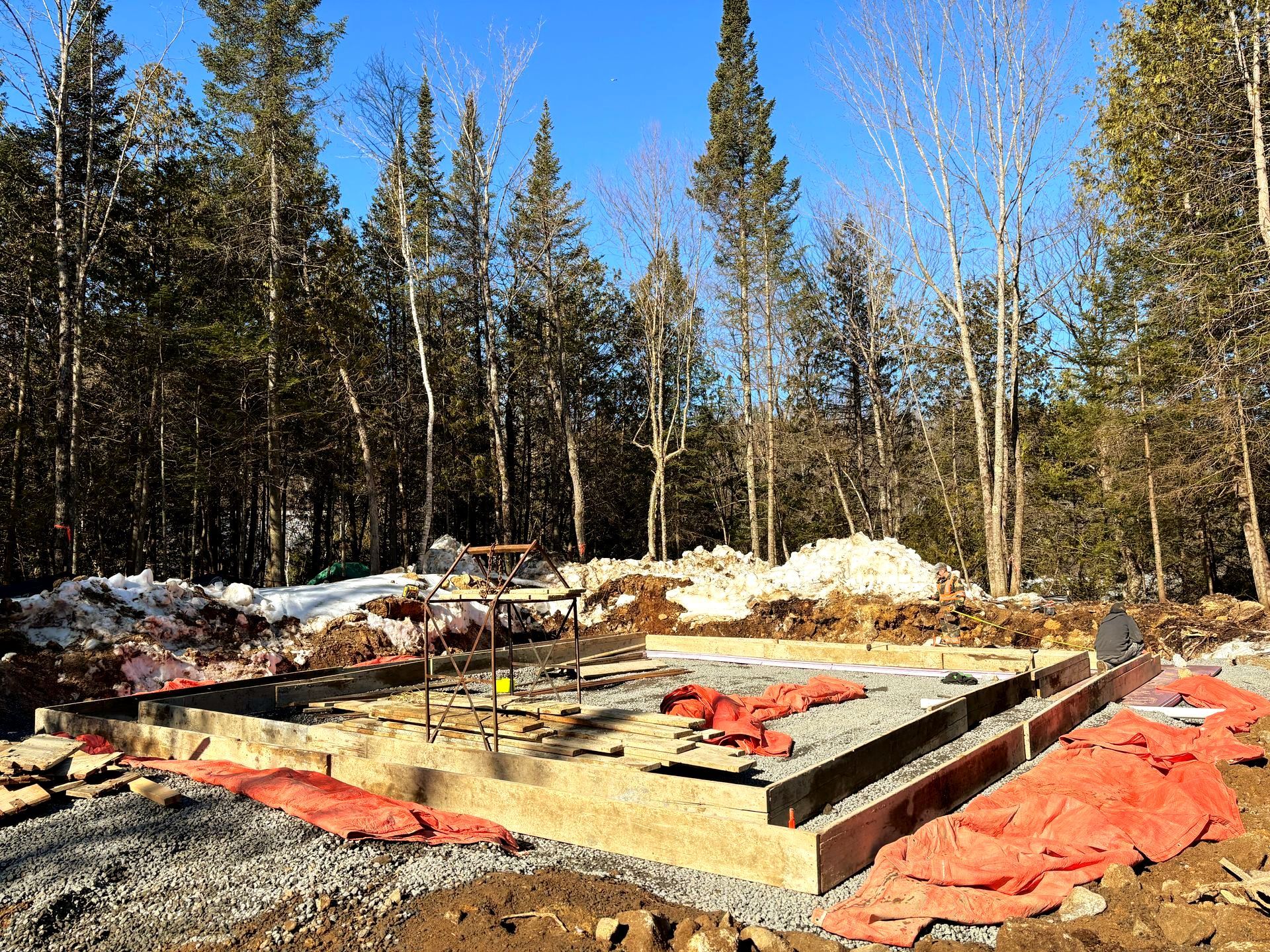 Construction site foundation in a snowy forest, with concrete forms and orange safety tarp under a blue sky
