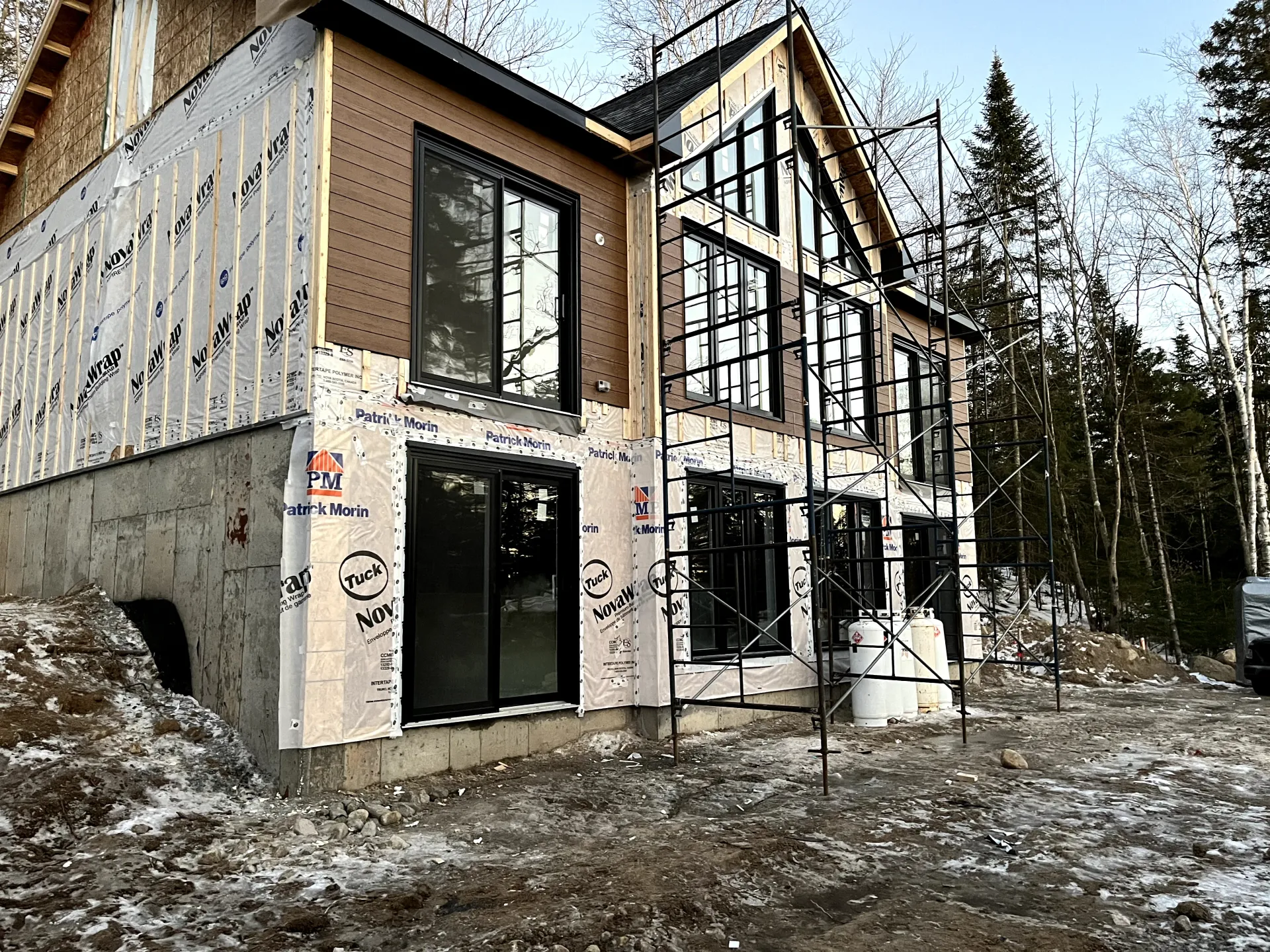 Modern house under construction with large windows, white wrap, and snowy forest surroundings
