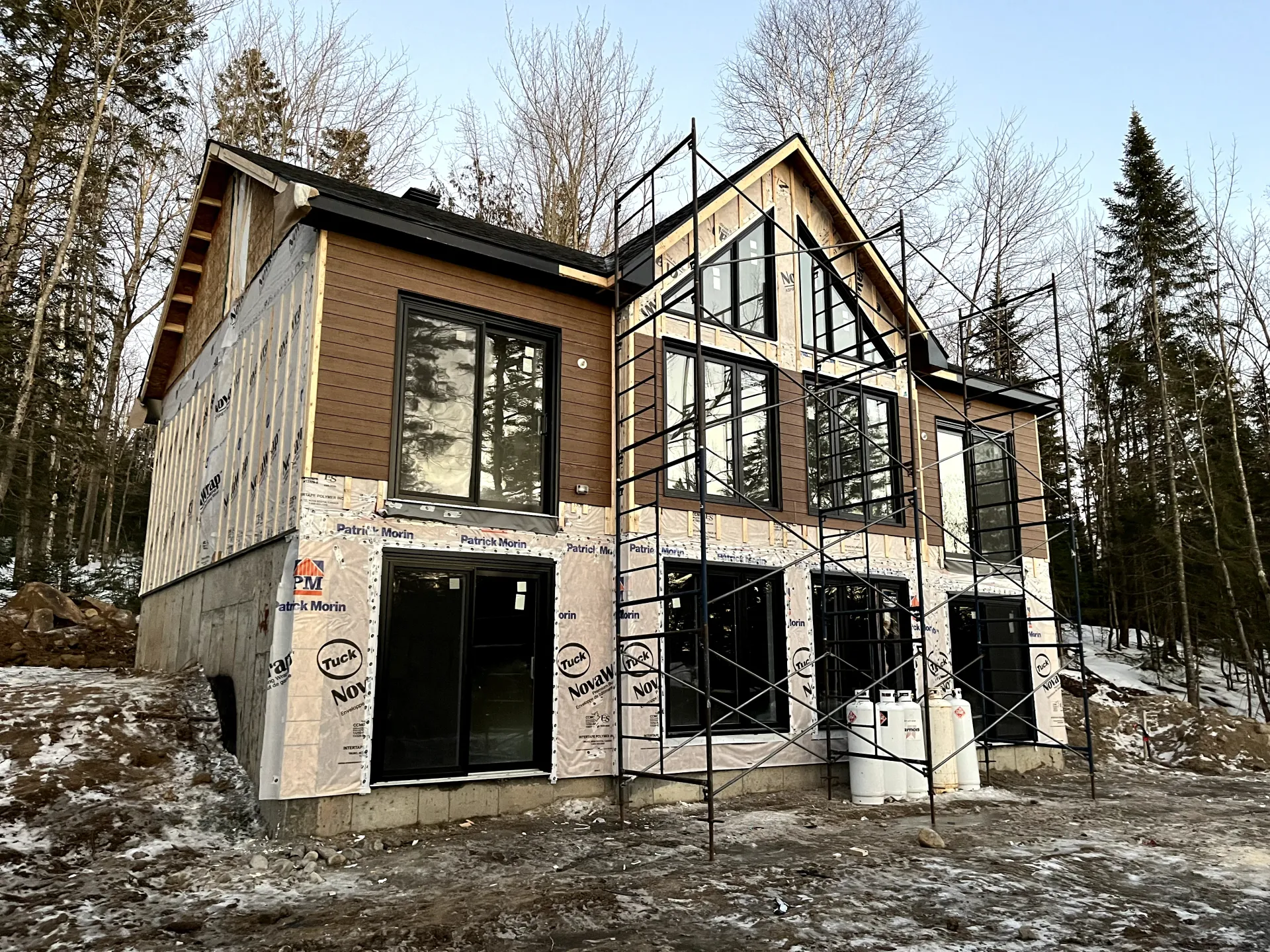 Modern two-story house under construction with exposed framing and snow on the ground, set among trees