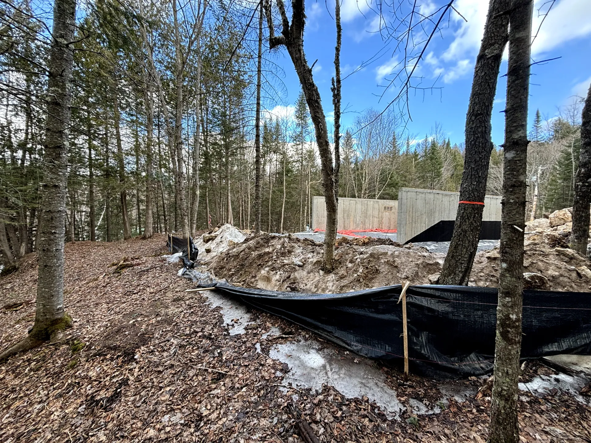 Wooded construction site with black silt fence, dirt piles, and bare trees under a blue sky