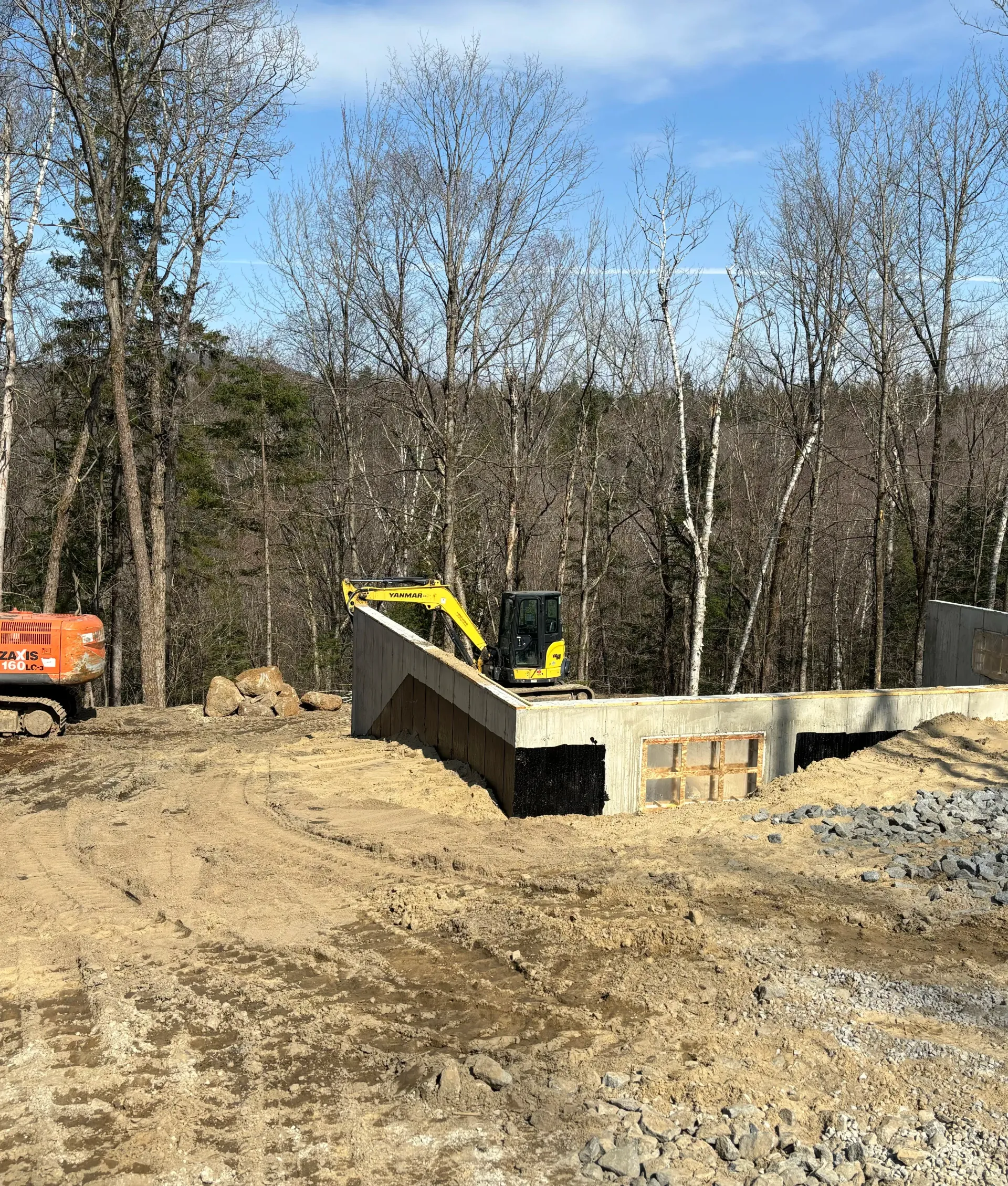 Bulldozer on a dirt construction site beside a concrete wall, with leafless trees in the background