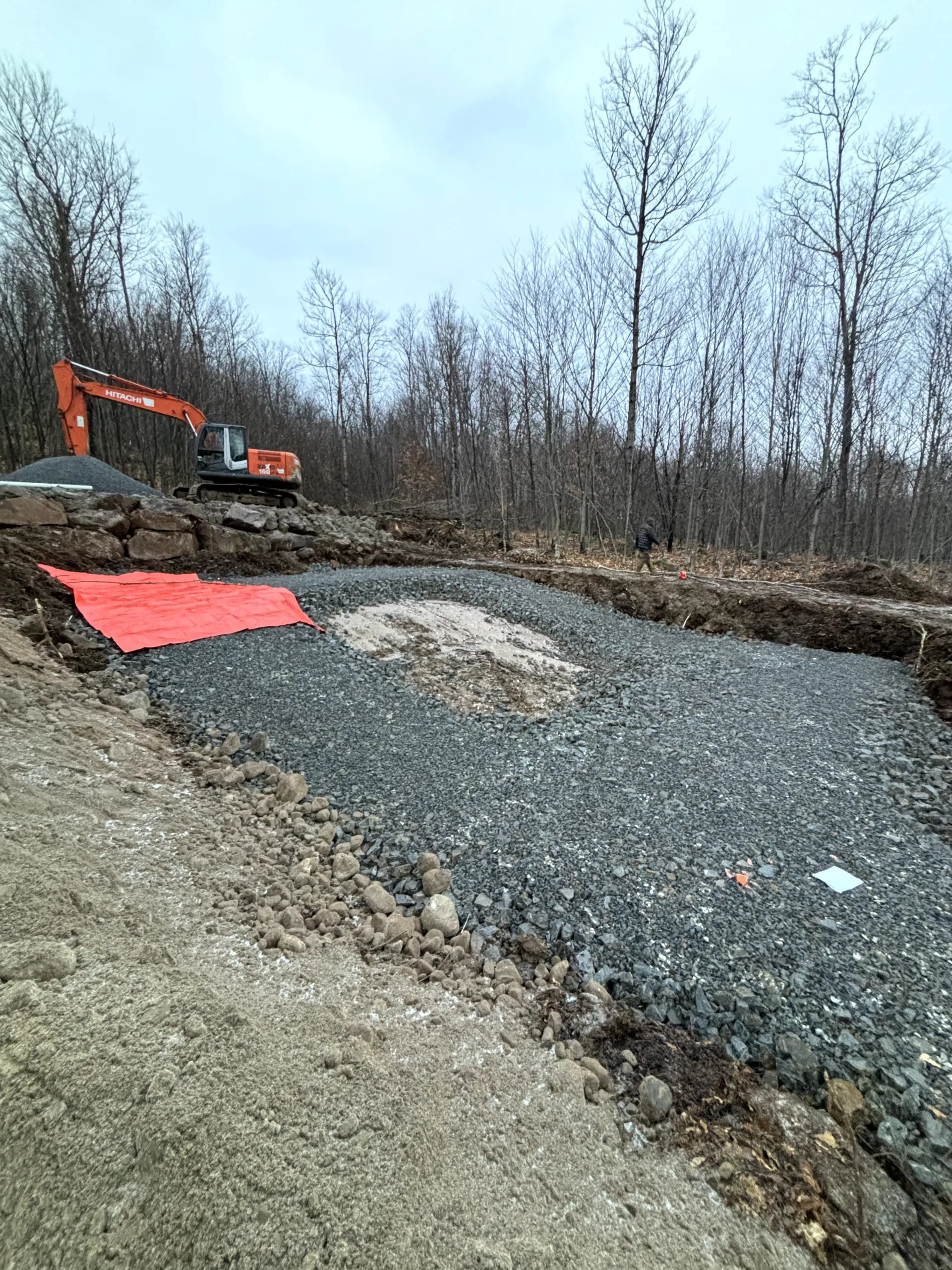 Excavator grading a gravel roadbed beside a wooded area, with red fabric and bare trees in view