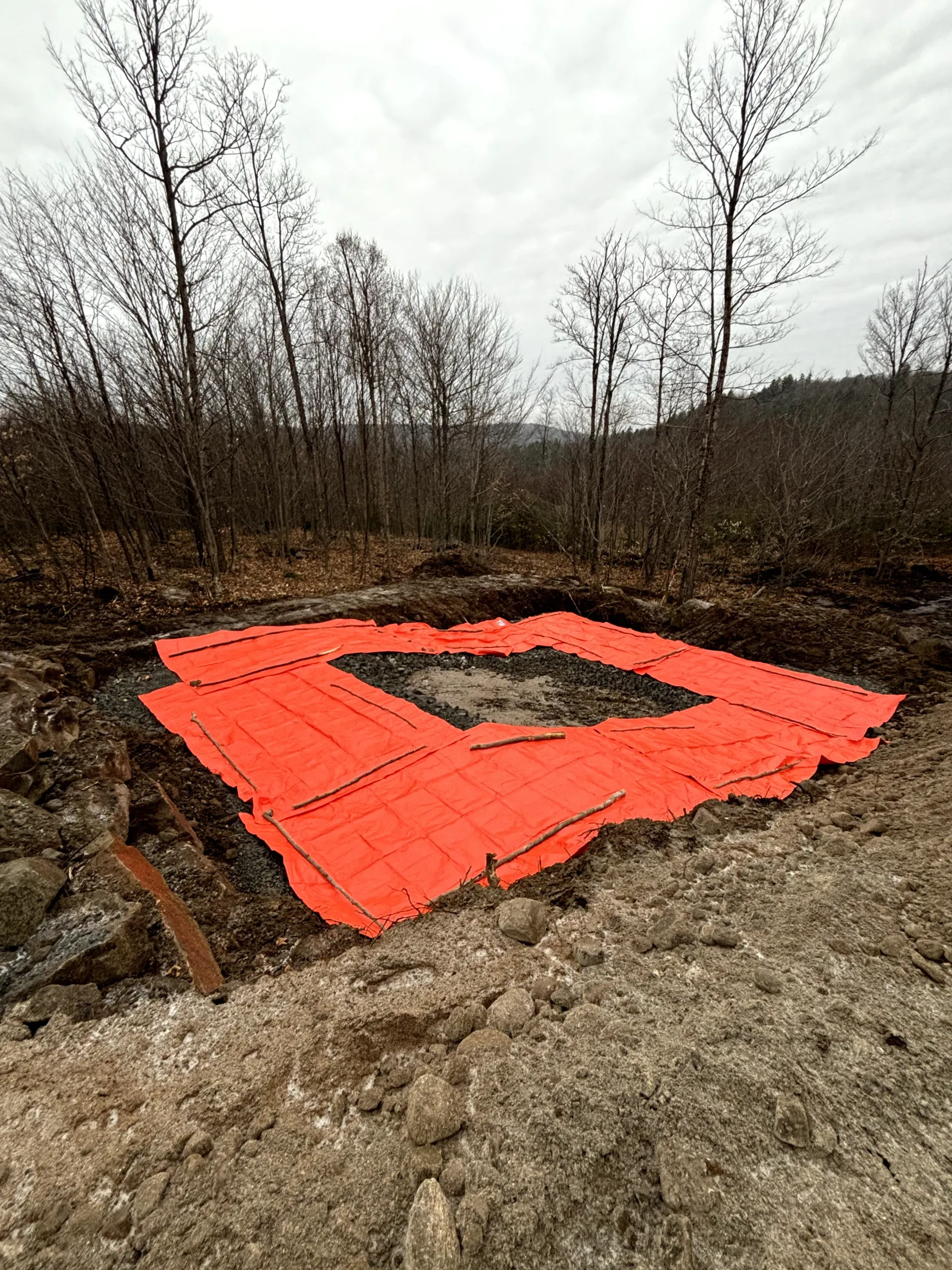 Bright orange containment mat around a square pit in a muddy, leafless wooded area
