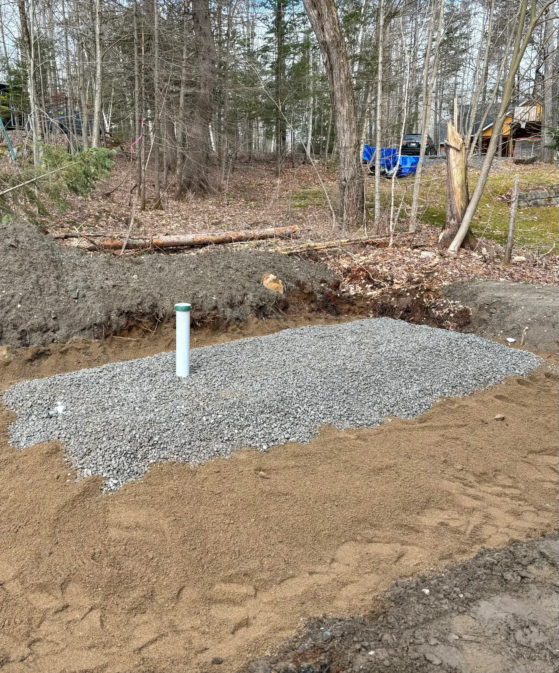 Construction site with gravel pad, white drainage pipes, and an excavator near a wooded area