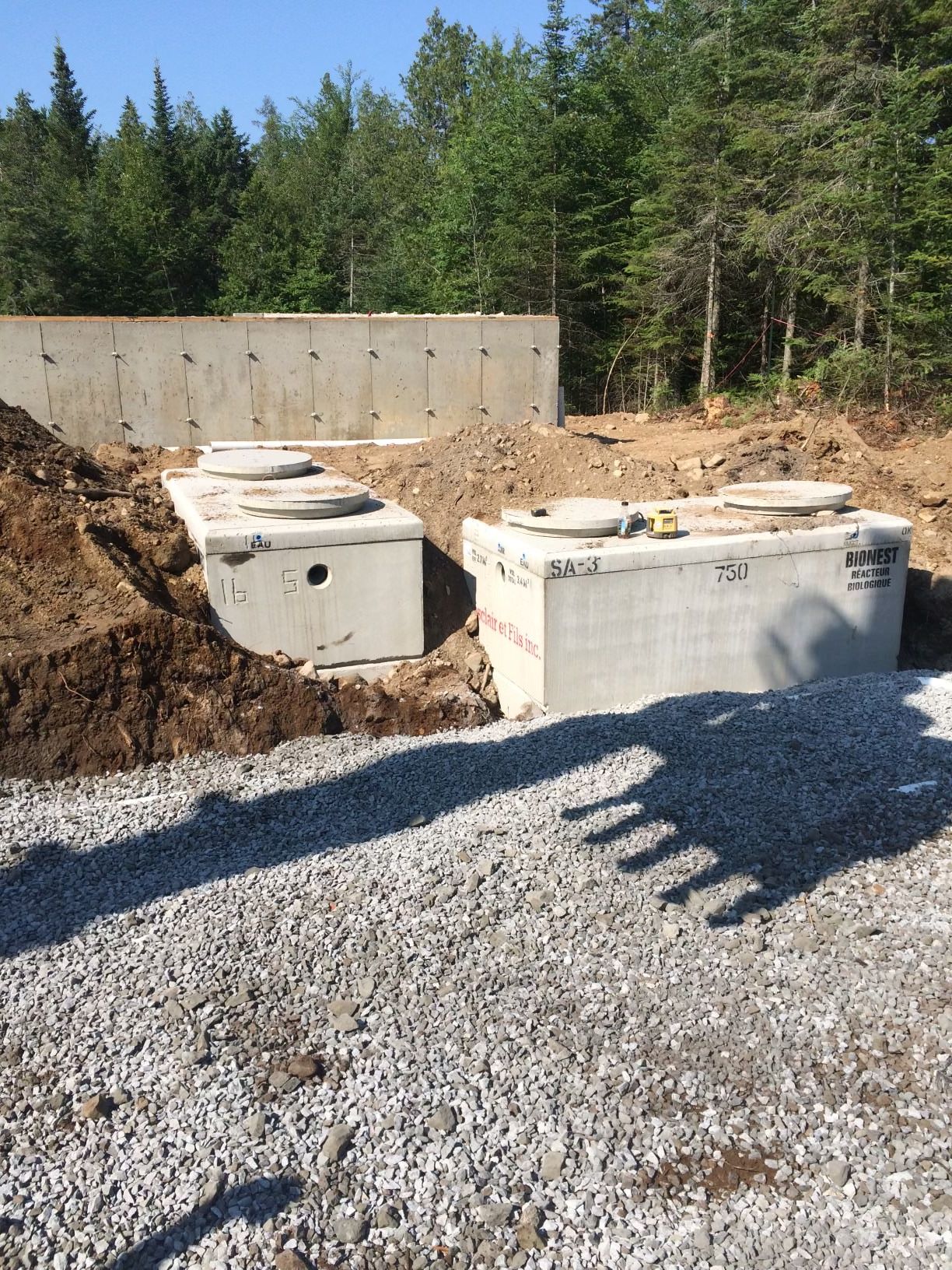 Concrete septic tanks and a retaining wall at a gravel construction site, with forest in the background