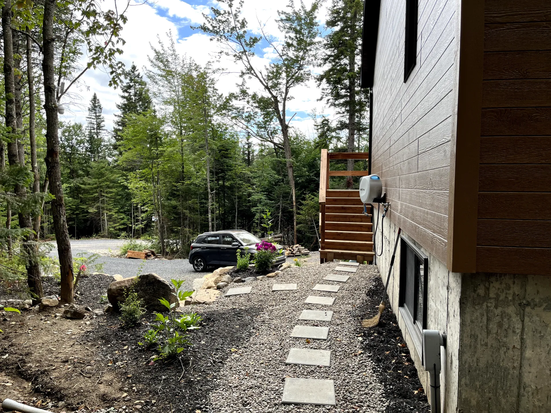 Gravel path beside a house leading to steps, with trees, rocks, and a parked car in a wooded yard.