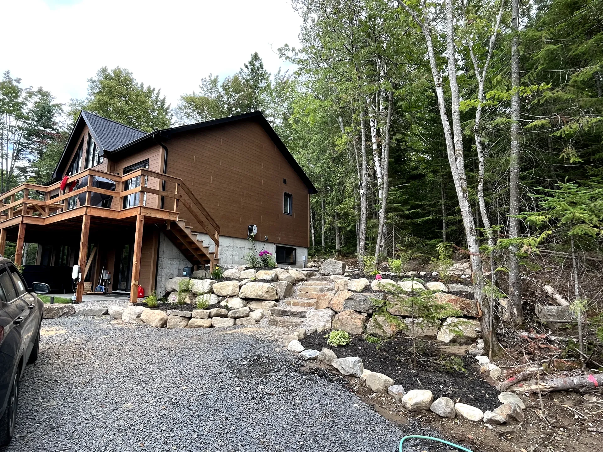 Rustic cabin with wooden deck beside gravel driveway and stone retaining wall in a wooded area