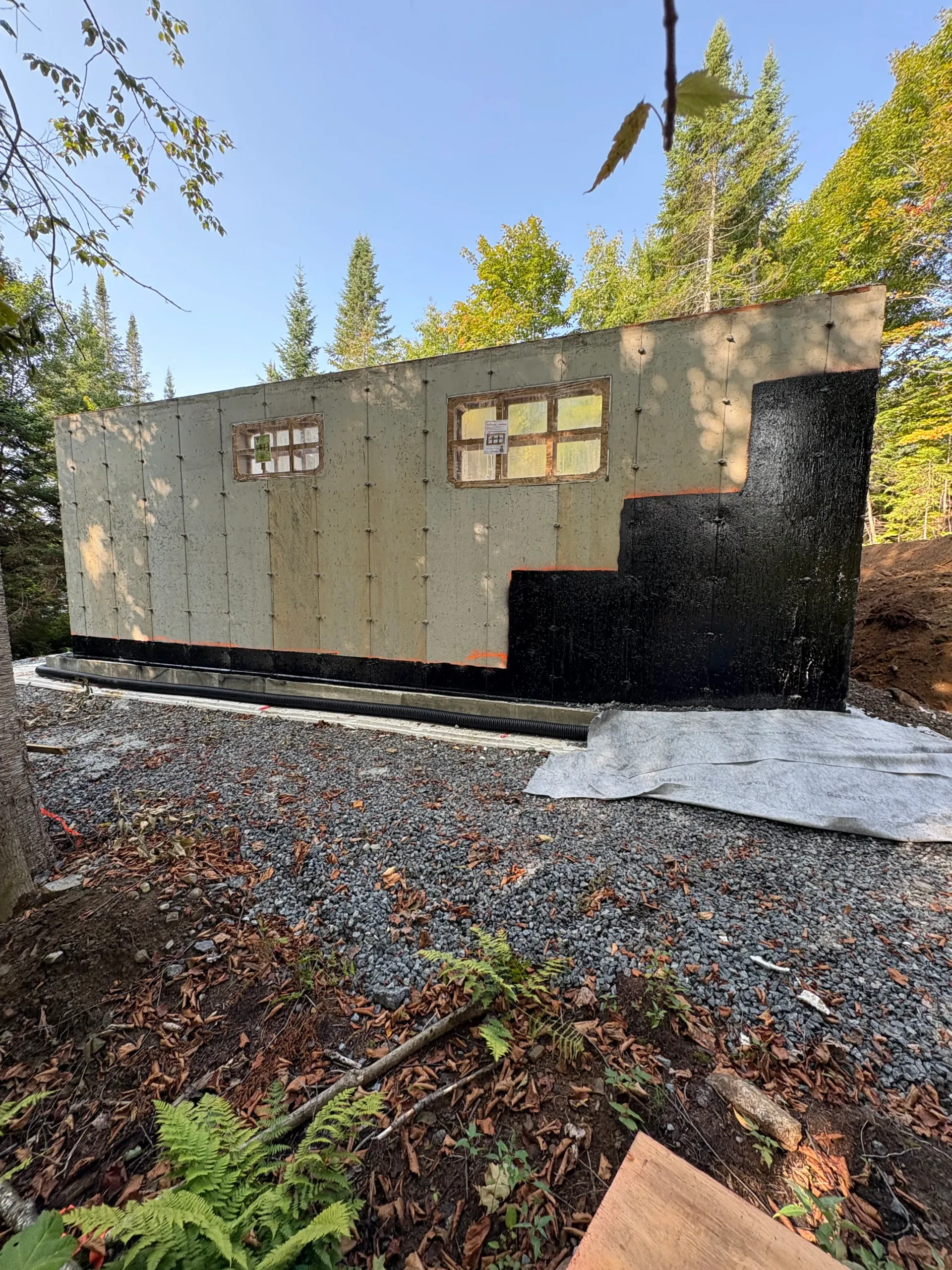 Small beige and black cabin in a wooded area, with a gravel foreground and bright sunlight.