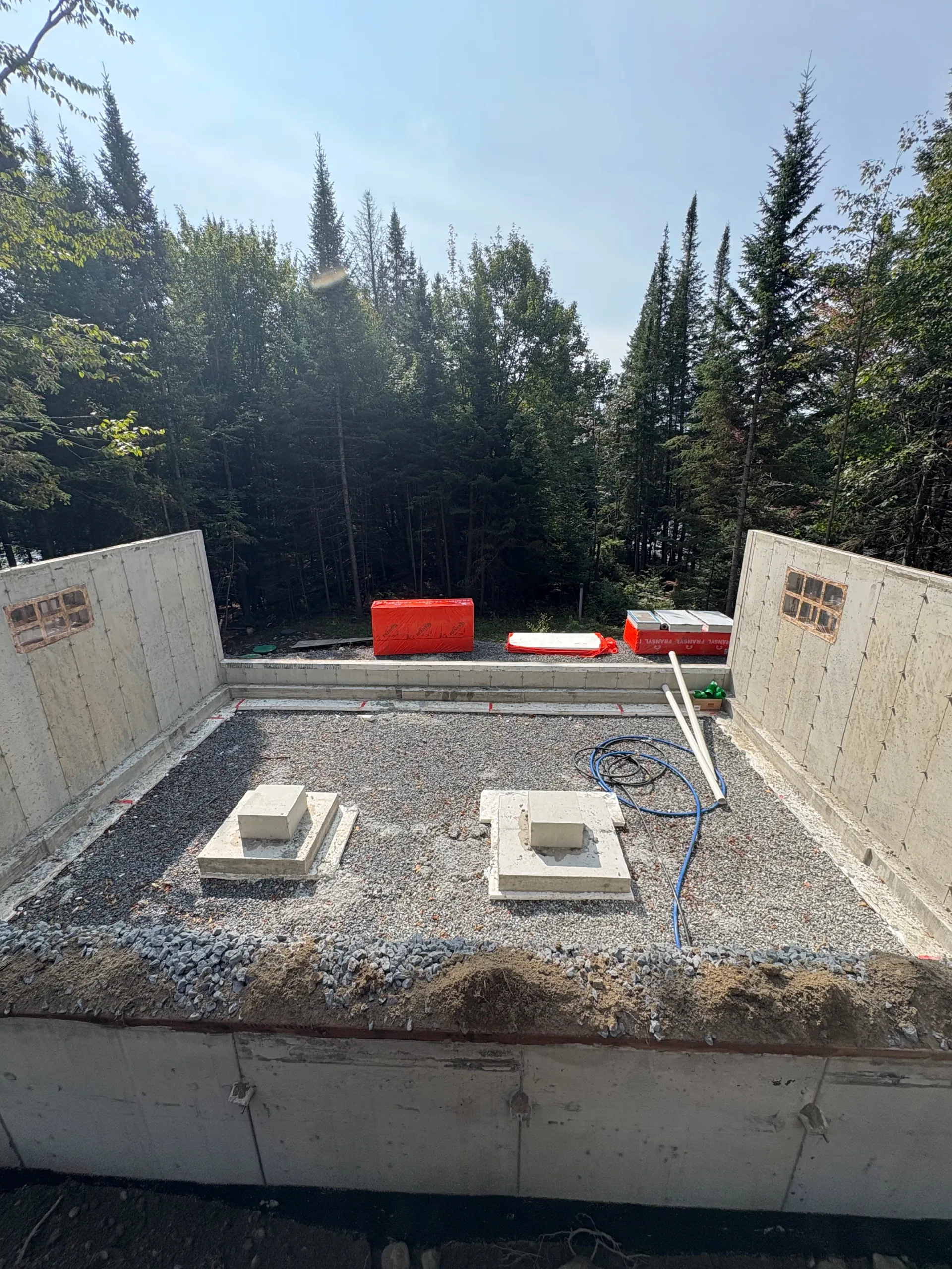 Concrete foundation with gravel, two square footings, and red formwork surrounded by trees under a bright sky