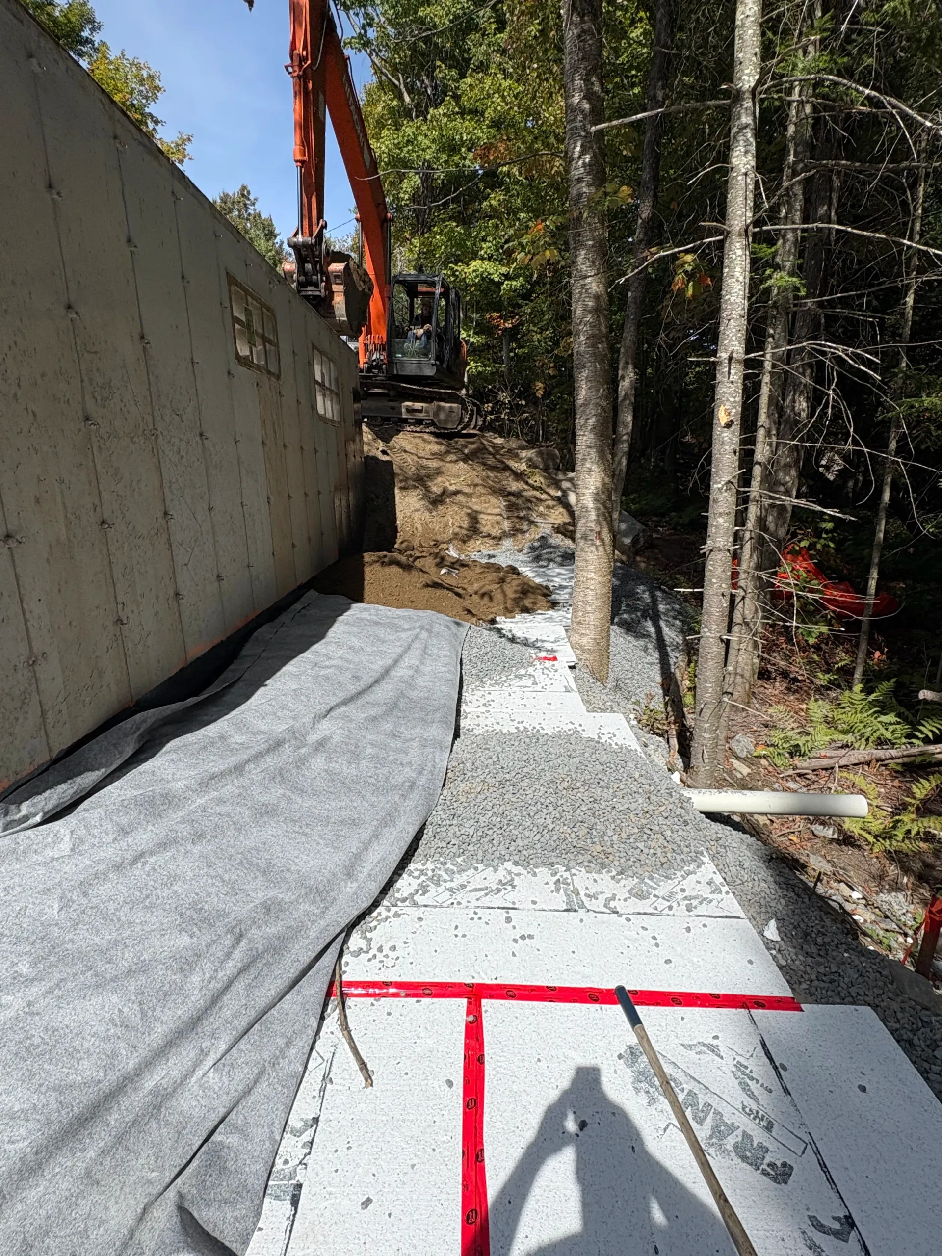 Construction site with concrete wall, gravel path, orange excavator, and red-marked ground in a wooded area