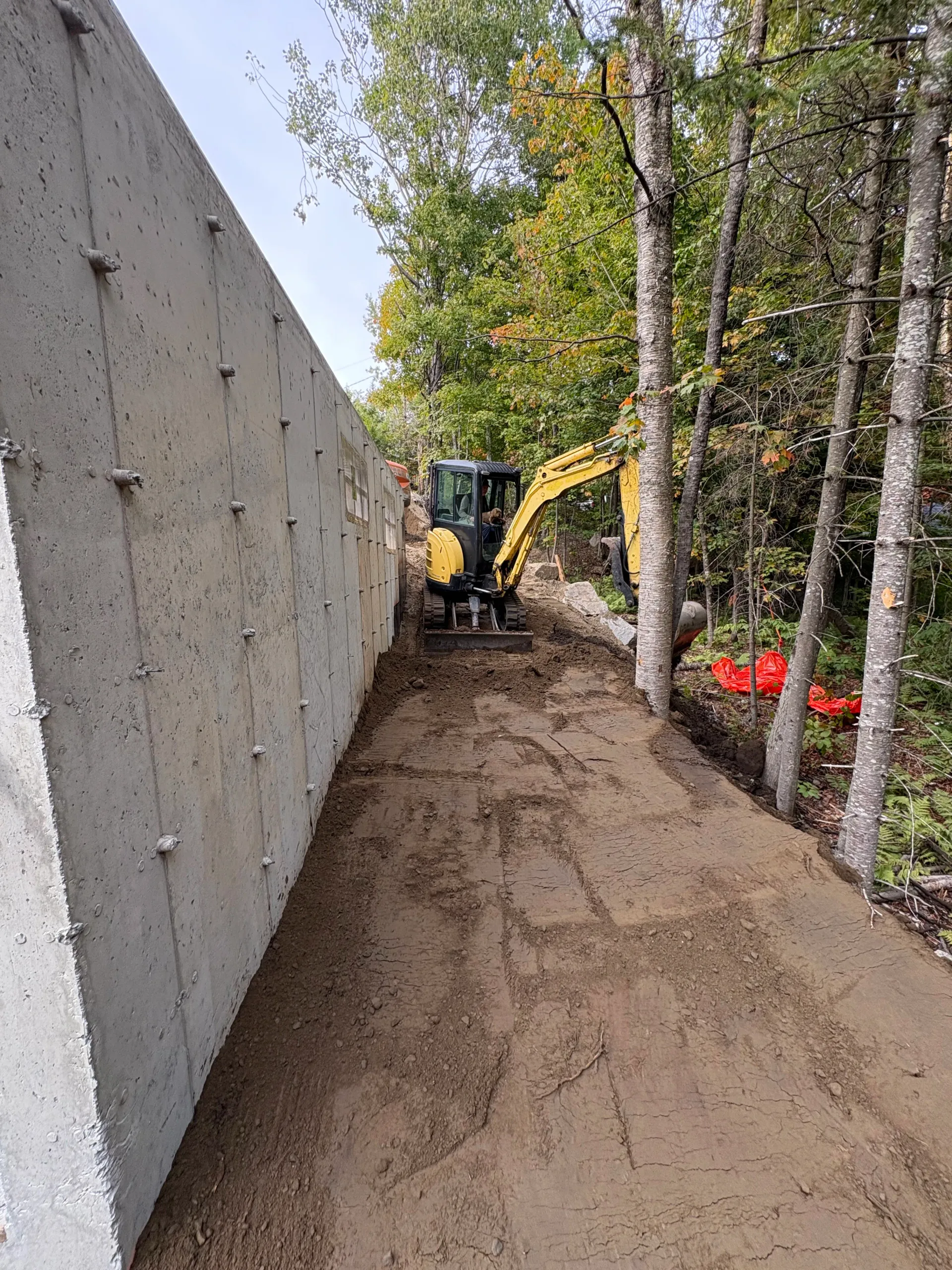 Excavator digging along a concrete wall beside trees, shaping a dirt path in a wooded area