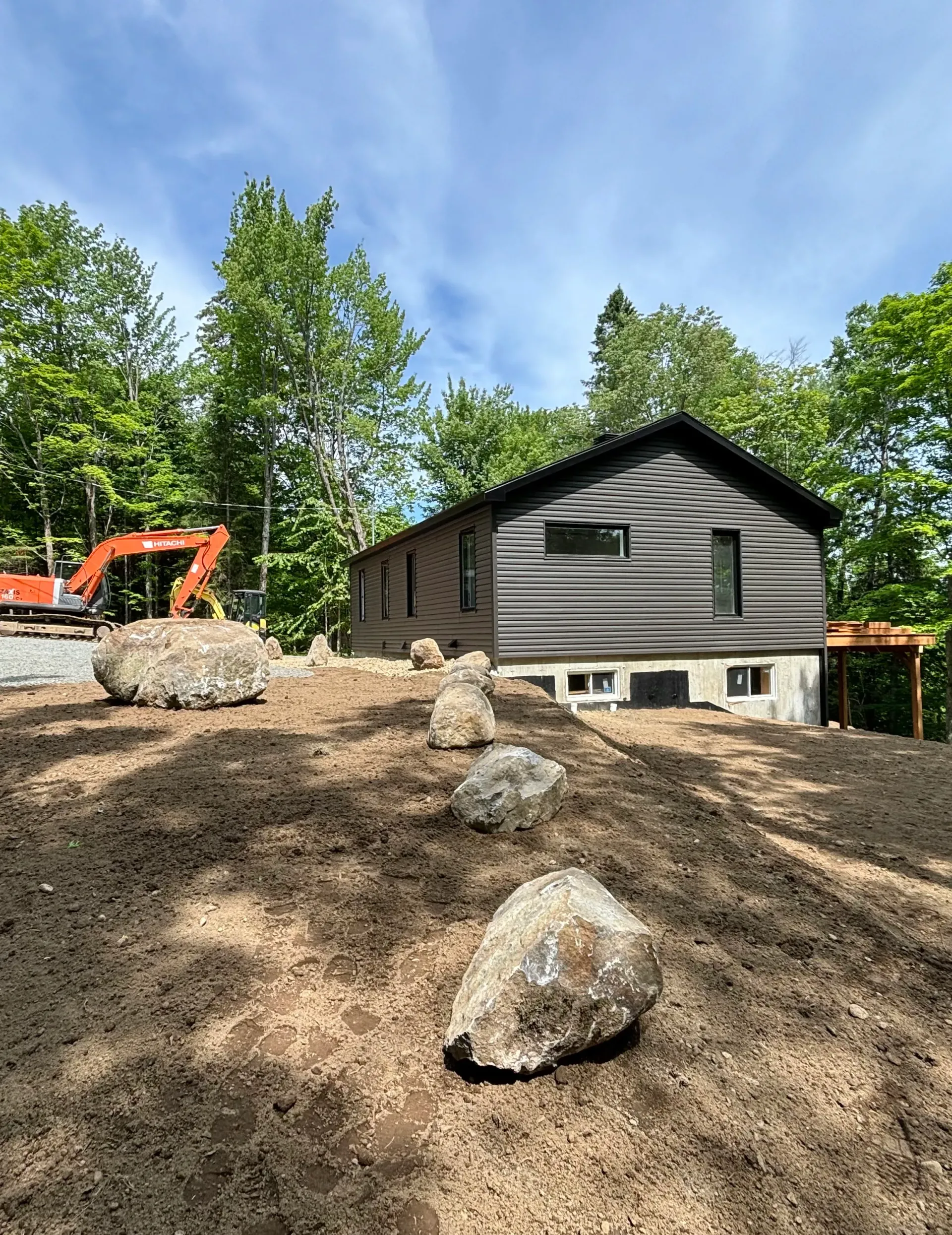 Gravel-covered yard with large rocks, a gray house, and an orange excavator beside trees under a blue sky