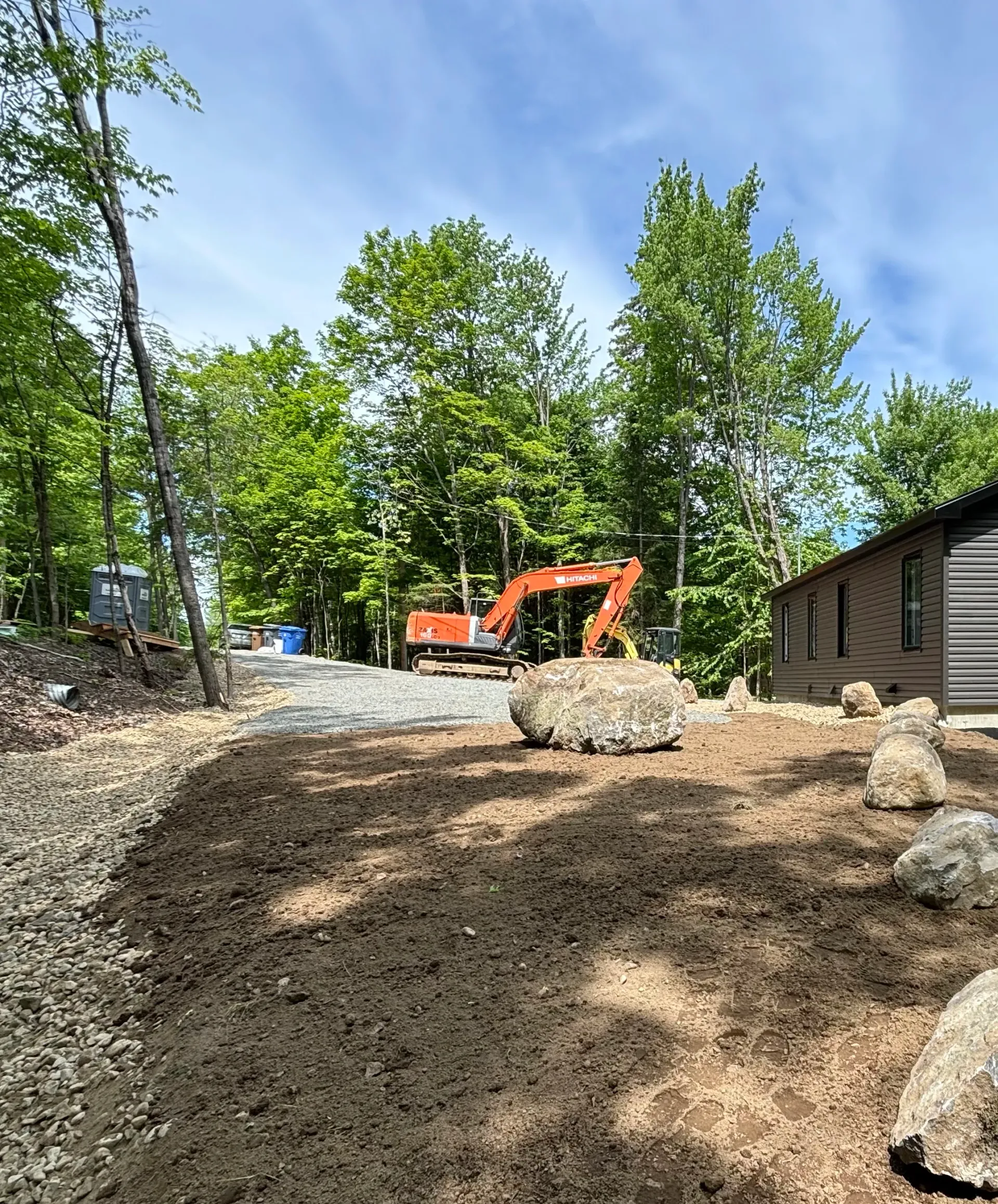 Orange excavator on a dirt lot beside a cabin in a wooded area under a blue sky