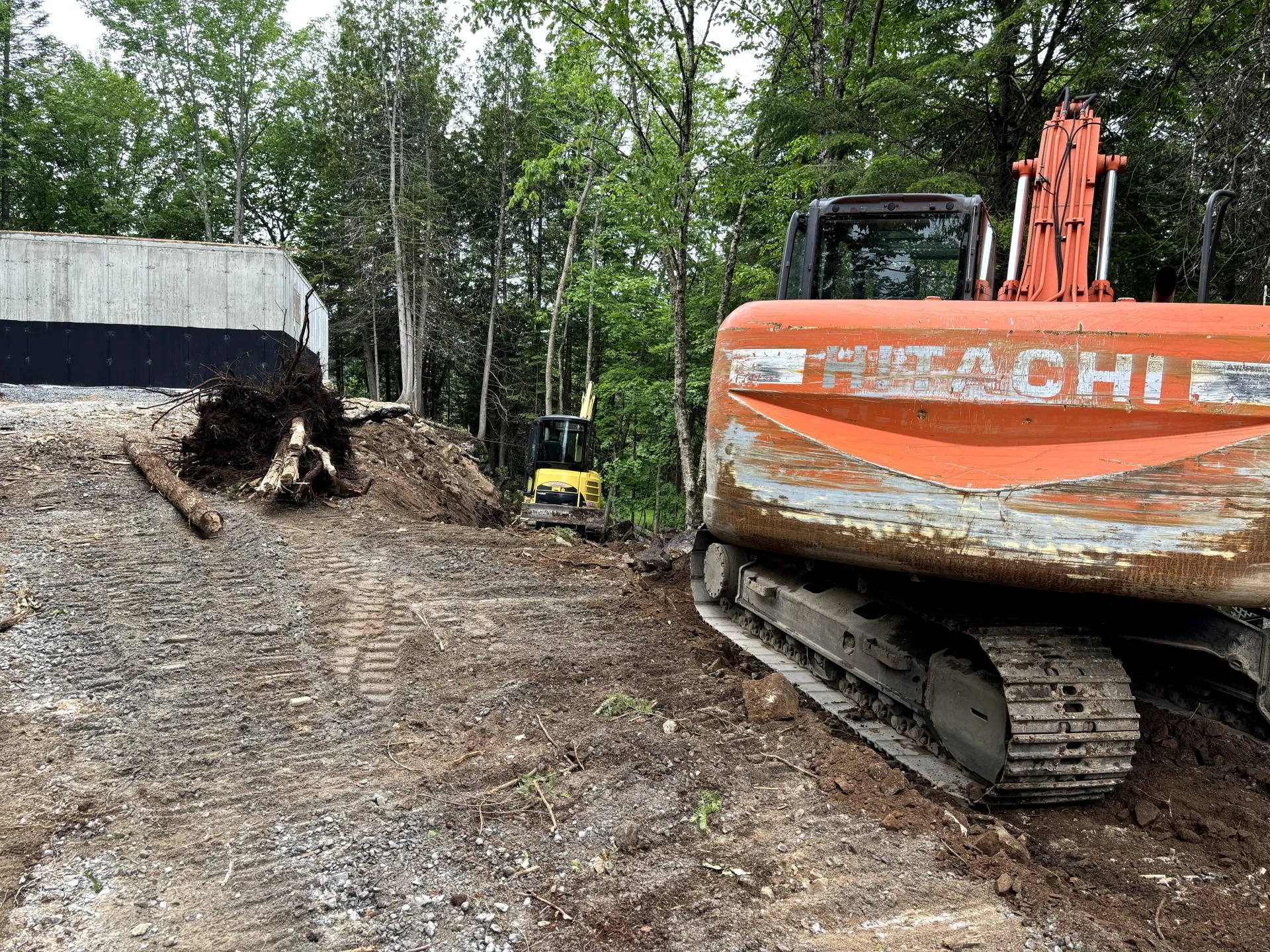 Orange excavator on a dirt mound at a wooded construction site, with a dumpster nearby.