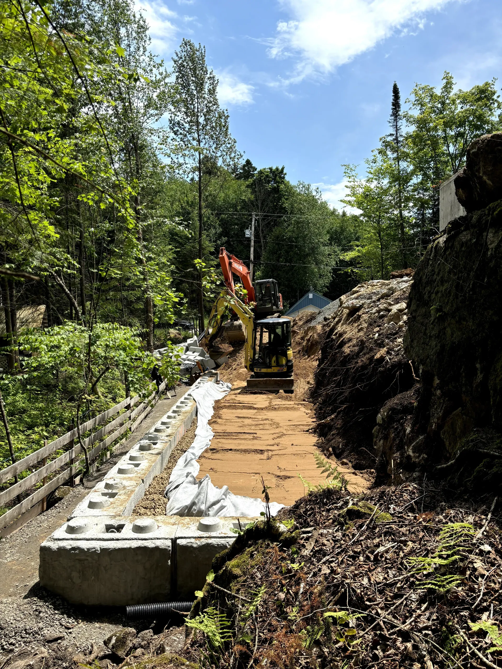 Construction worker paving a narrow path beside a rock wall in a lush, wooded area