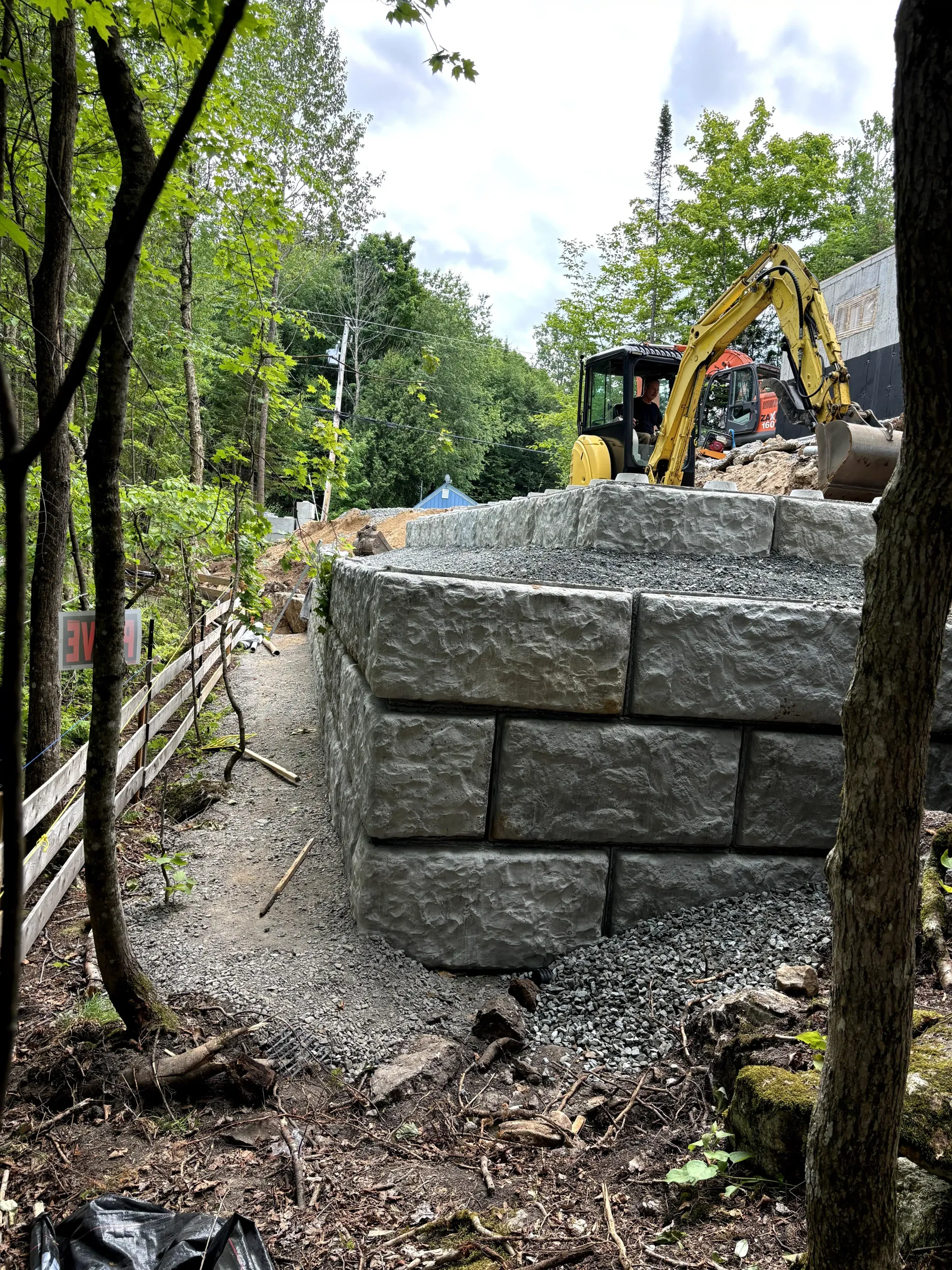 Stone retaining wall under construction beside a wooded path, with an excavator in the background