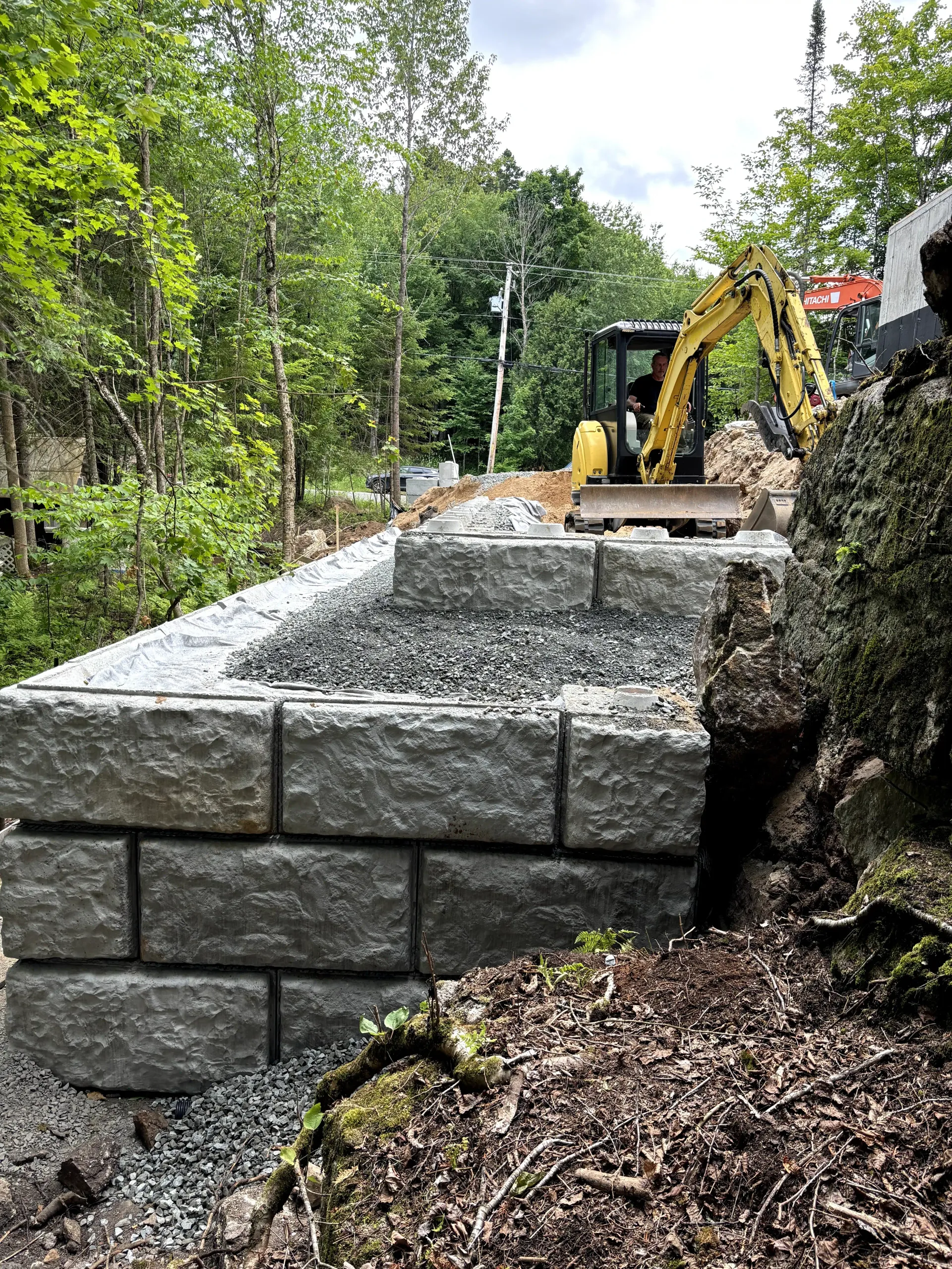 Stone retaining wall under construction beside a wooded path, with an excavator in the background